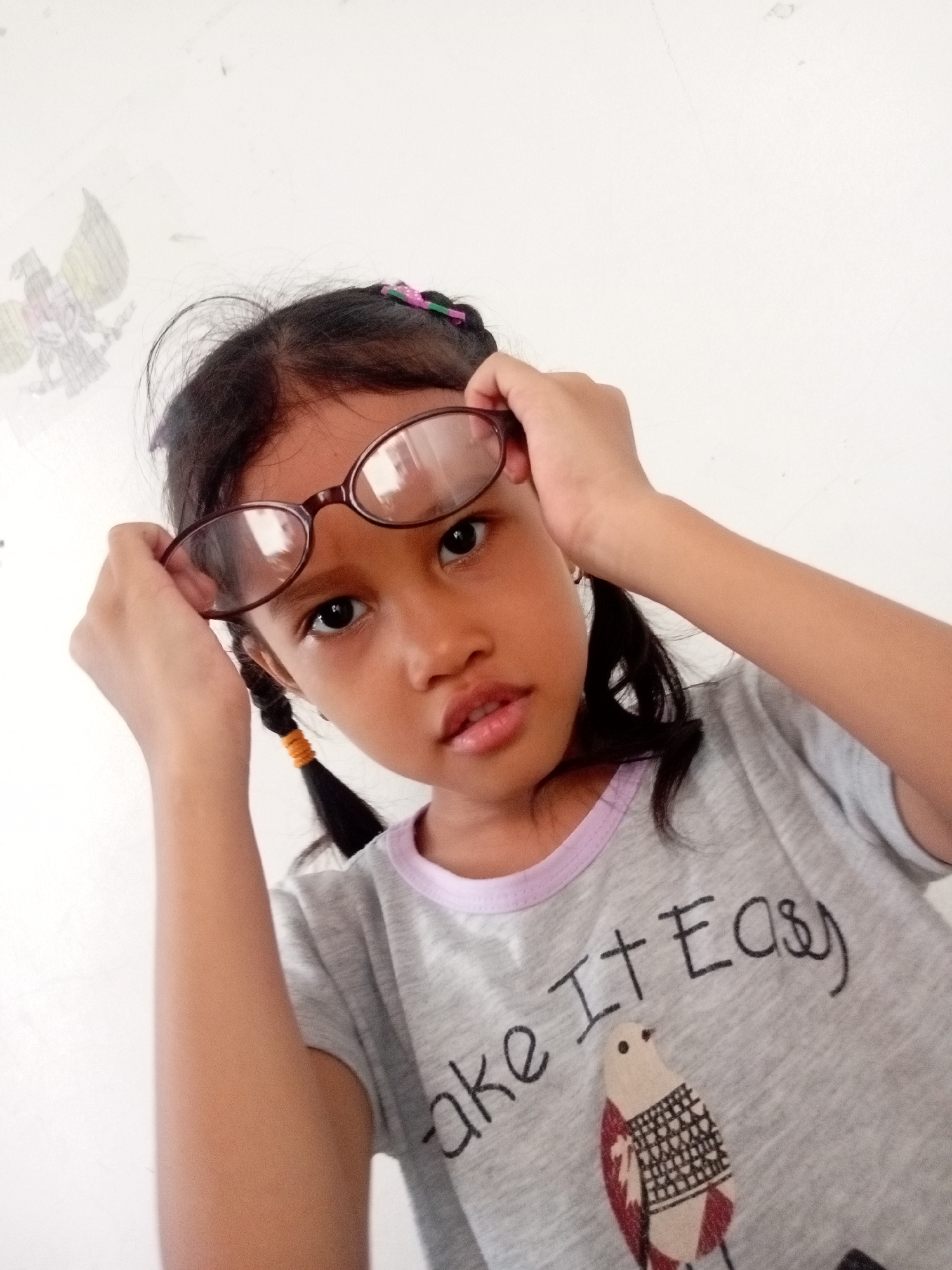 A young girl with dark hair in pigtails, wearing glasses and a gray t-shirt with a bird graphic and the words "Take It Easy," is holding her glasses up. She is positioned in front of a white wall with a drawing of a bird on the left side. The image appears to be taken indoors, likely in Batam, Indonesia, given the drawing which resembles the Indonesian coat of arms (Garuda Pancasila).