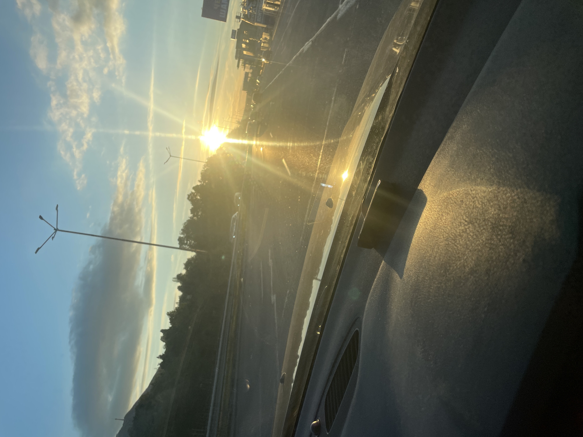 The image depicts a view from inside a vehicle, likely a car, looking out through the windshield. The perspective is low and slightly angled upward. The dashboard of the car is visible in the foreground, dark in tone. Outside the vehicle, the sun is low on the horizon, creating a bright glare and multiple light flares that partially obscure the view. In the middle ground, there are trees and possibly some buildings. A road is visible, with streetlights lining the side. The sky is a mixture of blue and yellow, with some clouds present. The location is a city, as evidenced by the presence of buildings and streetlights, but the specific city is unknown. There are no people visible in the image. The predominant action is driving, implied by the interior view from a vehicle.