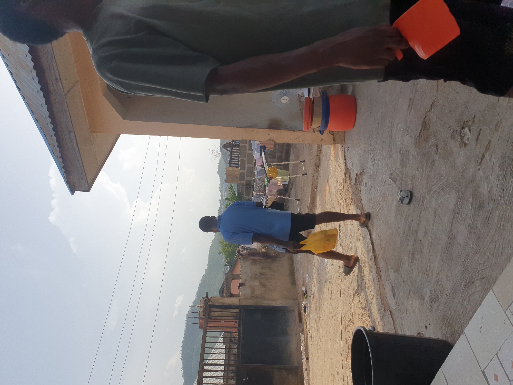 The image captures an outdoor scene in Lokoja, Nigeria. A man in a blue shirt and dark shorts is walking away from the camera. He is carrying a yellow object, possibly a bag or container. The ground appears to be concrete or paved, and there are buildings in the background. A clothesline with drying laundry is visible further away. A metal gate is present towards the bottom of the image. Two plastic buckets and an electric fan are seen along the wall of the house. Another person in a green shirt stands near the camera. The sky is partly cloudy.