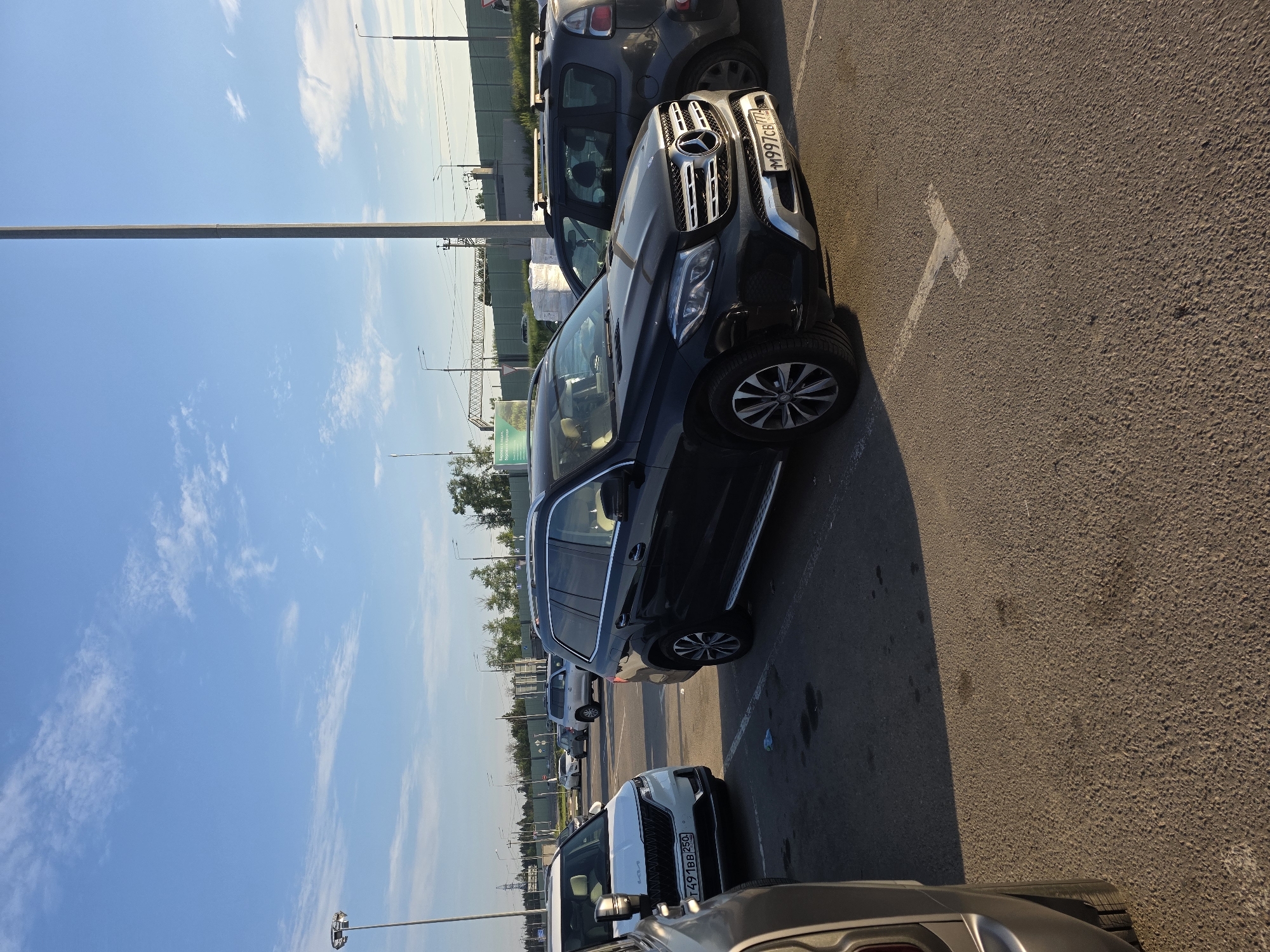 A wide shot captures a parking lot under a clear blue sky with scattered clouds. In the foreground, a black Mercedes-Benz SUV is parked. To its left, a white Kia minivan is visible, and further left, the rear section of a grey SUV occupies the frame. In the background, several other vehicles are parked, including a dark grey hatchback. A tall, thin lamppost stands prominently on the left side of the image, and another lamppost is visible further back on the right. Beyond the parking lot, a green sound barrier is present, with trees and some industrial structures in the distance. The scene appears to be an outdoor parking area with marked parking spaces.