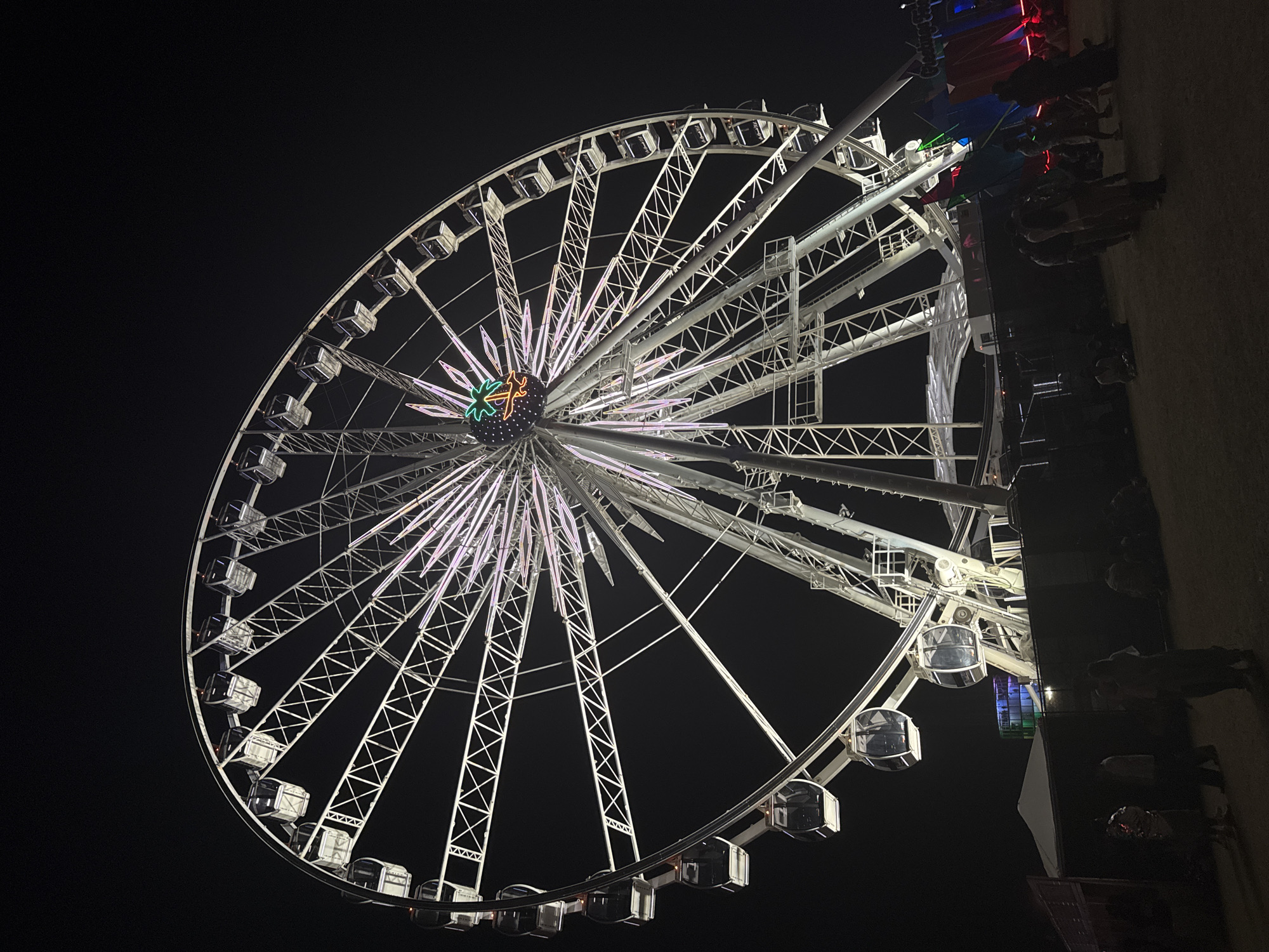 The image shows a brightly lit Ferris wheel at night. The wheel is white with numerous enclosed passenger cars. The center of the wheel has a colorful design, possibly a logo. The background is a dark night sky. To the right of the wheel, a crowd of people is visible, suggesting a fair or festival setting. The location is Indio, California.
