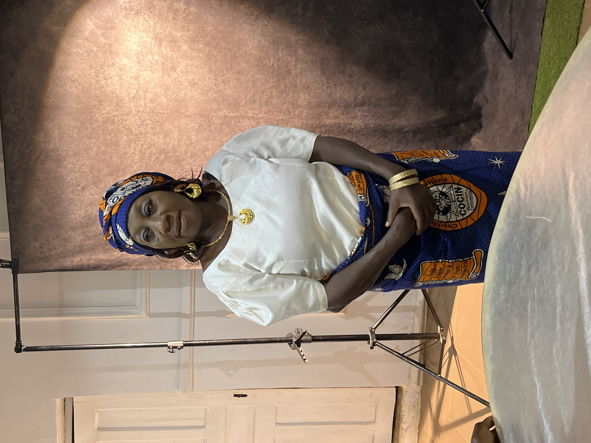 A woman stands in a studio, facing forward. She has dark skin and dark hair, styled under a blue and orange headwrap. She wears a white, puffed-sleeve blouse and a blue skirt with an orange and white pattern. Gold earrings, a gold necklace with a large pendant, and two gold bracelets adorn her. Her hands are clasped in front of her.
To the left of the woman, a white door with a dark handle is visible, set into a white wall. A metal tripod with a light attached stands between the woman and the door. The background consists of a mottled brown backdrop. A large, light-colored, curved object occupies the foreground, partially obscuring the bottom of the image. The floor is tiled and partially covered with a green, mat-like material.
