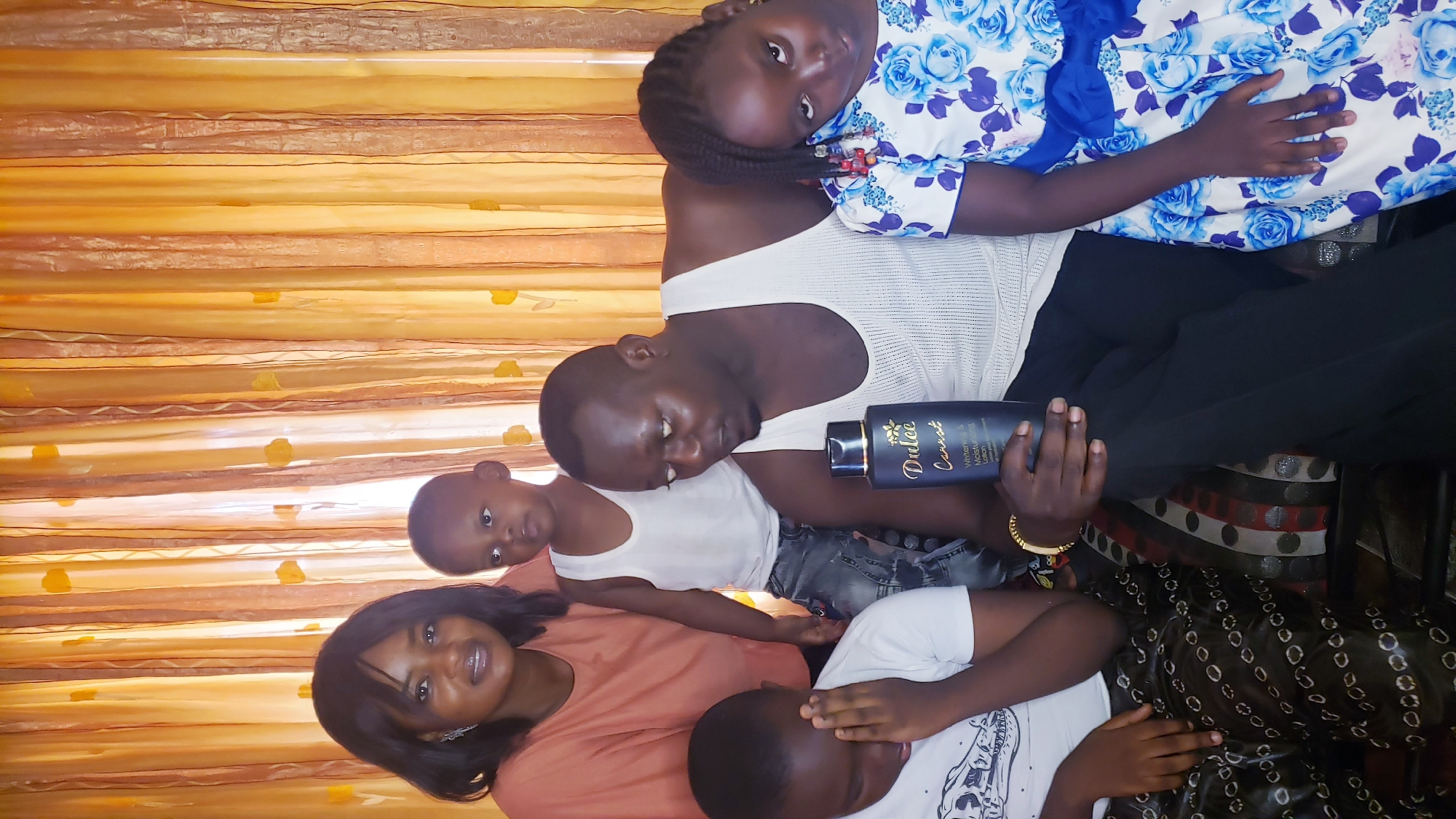 The image depicts a family of five indoors, posing for a photo. The setting includes a backdrop of orange drapes and patterned furniture, possibly a couch or chair. A woman is in the foreground wearing a peach-colored top. A man in a white tank top holds a bottle of "Dulce Carrot" whitening and moisturizing lotion, with a young girl wearing a blue floral dress resting on his shoulder. Two younger boys are also in the frame, one in a white tank top and the other in a white t-shirt, partially covering his face with his hand. The family appears to be situated in Sabon Tasha, Nigeria.