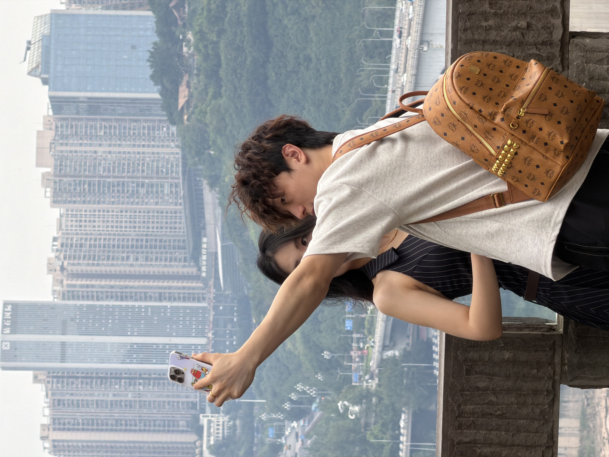 A young couple is taking a selfie in front of a dense urban skyline in Yuzhong District, China. The man in the foreground, wearing a light grey t-shirt and a brown MCM backpack, is holding a smartphone with a cartoon case to capture the image. His right arm is extended, holding the phone, while his left arm is down. The woman, partially visible behind him, wears a black and white pinstripe dress and has long dark hair. She is looking towards the camera. The background is dominated by numerous high-rise buildings, some appearing to be residential apartments, clustered on hillsides. A large body of water is visible in the distance below the buildings, along with roads and bridges. The overall scene depicts a moment of tourism and connection amidst a vast city.
