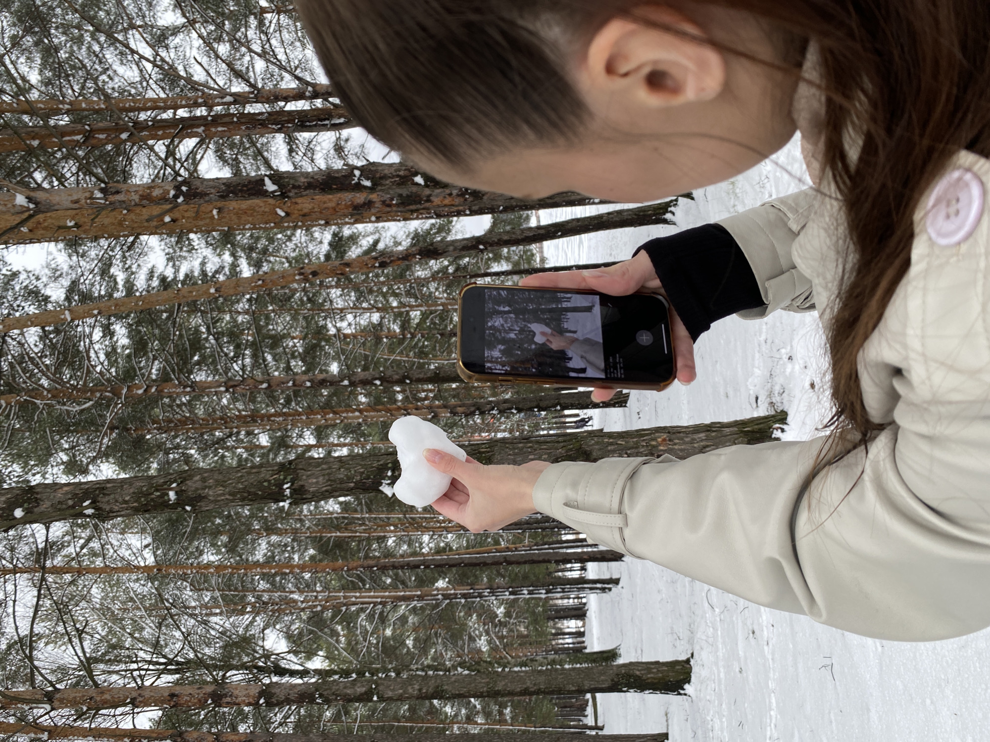 A woman is taking a picture of a snowball with her phone in a snowy forest. The forest has many tall trees. The phone is held in her left hand. She is wearing a light-colored jacket and a black undershirt. Her hair is pulled back. The location is the city of Chaikovsky, Russia.