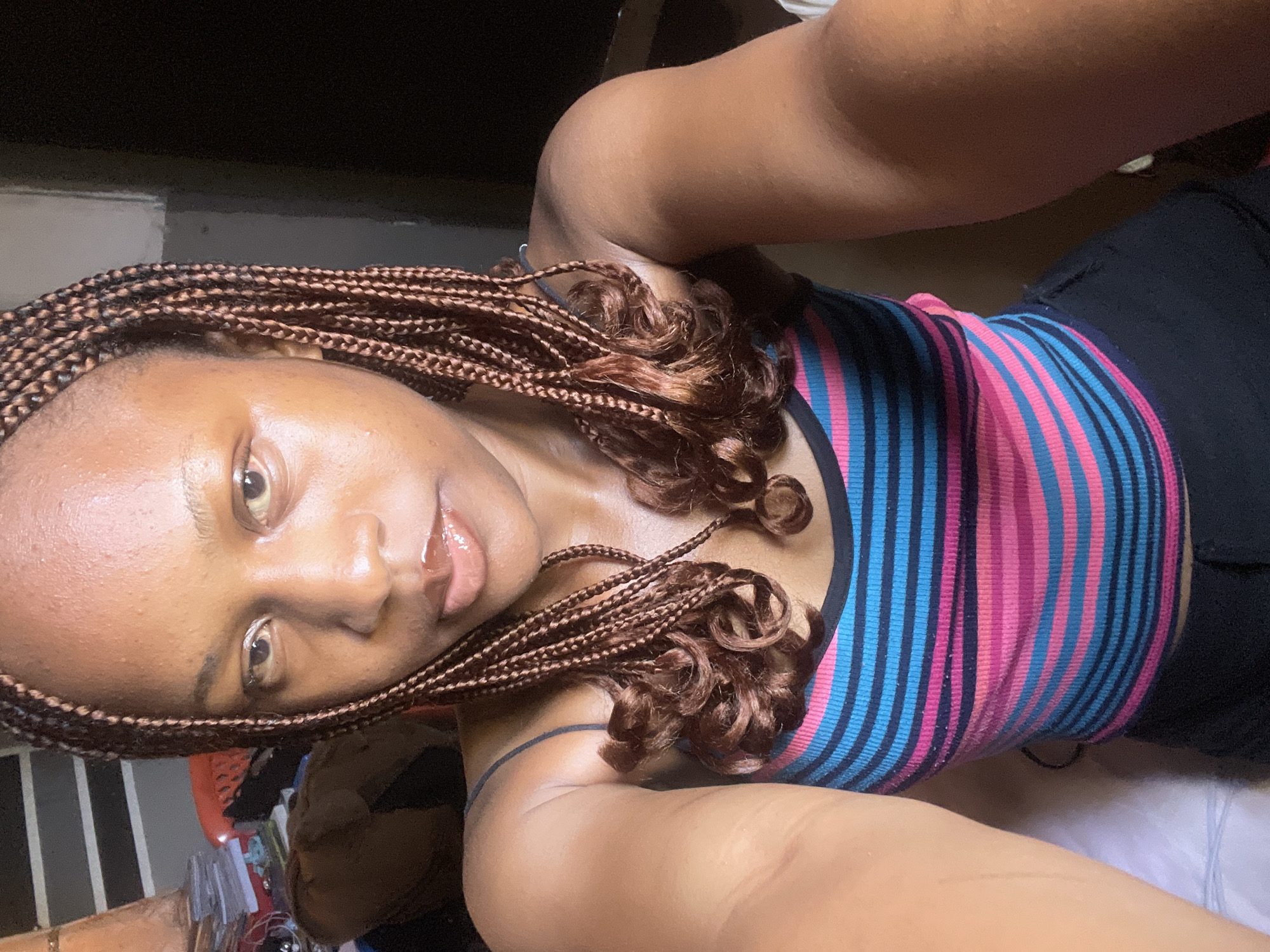 The image is a close-up, waist-up selfie of a young Black woman with braided hair. She is wearing a striped tank top with pink, blue, and black horizontal lines, and dark-colored pants. Her face is turned towards the camera, and she has a neutral expression. The background appears to be an indoor setting with some shelves and stacked items on the left side, and a dark wall on the right. The lighting suggests it is daytime, possibly with natural light from a window. The image does not provide any information that would suggest it was taken in Jos, Nigeria.