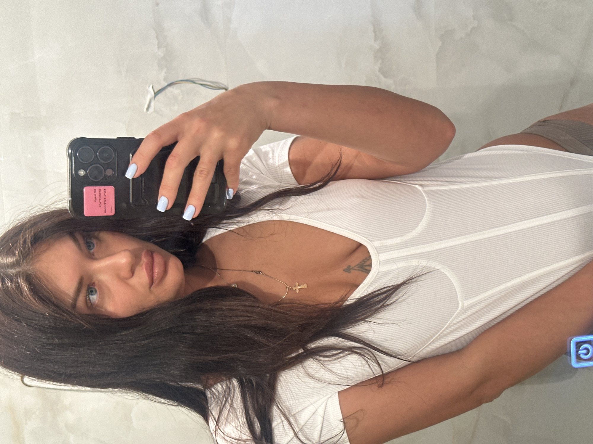 A young woman is featured in a mirror selfie, holding a black smartphone with a triple-lens camera array and a pink sticker on its back. Her fingernails are painted a light blue. She has long, dark brown hair, blue eyes, and full, naturally colored lips. She wears a form-fitting, white ribbed top with structural detailing around the bust. Two thin gold chains adorn her neck, one featuring a small cross pendant. A faint tattoo outline is visible on her upper chest/shoulder area. Below the white top, a glimpse of a darker, ribbed garment is visible. The background consists of a light-colored, possibly textured wall or reflective surface, with some exposed wires visible towards the top. The woman maintains a neutral expression, looking directly into the phone's camera. The pink sticker on the phone case displays Cyrillic text, reading "БЕЛОРУССКИИ" (Belarusian) and "ГОСУДАРСТВЕННЫЙ" (State).