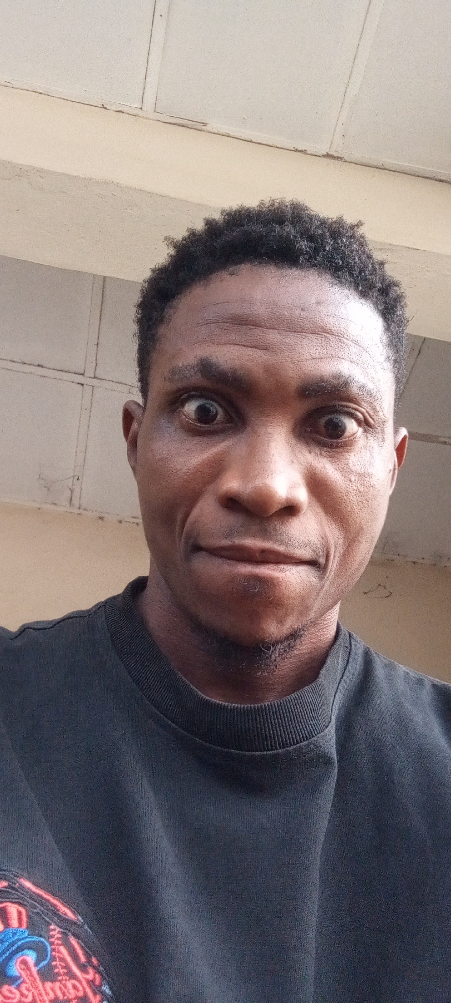 A close-up shot of a dark-skinned man with short, curly black hair. He's wearing a black t-shirt and has a slight goatee. His eyes are wide open, giving him a surprised expression. The background suggests an indoor setting with a light-colored wall and ceiling, possibly in Adankolo, Nigeria.