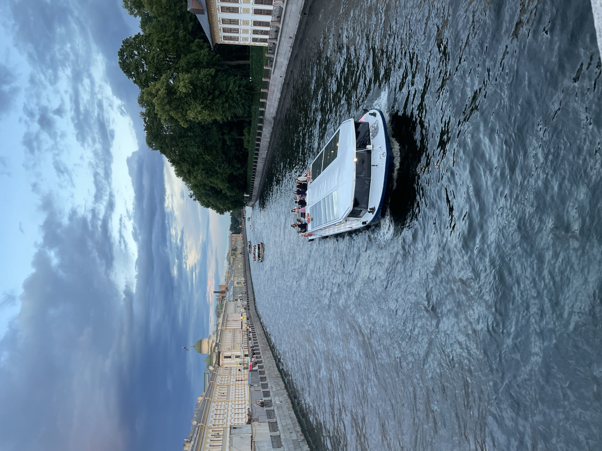 The image depicts a wide, dark river flowing alongside a city embankment. On the river, a white tour boat with a glass enclosure carries several passengers, appearing to be on a sightseeing excursion. The boat leaves a white wake as it moves. In the background, a long, ornate building with multiple windows and a light-colored facade lines the embankment. Lush green trees grow along the edge of the embankment, casting shadows on the water. Further in the distance, more buildings and a dome with a flag can be seen. The sky is dramatic, filled with a mix of dark, heavy clouds and lighter, wispy ones, suggesting either early morning or late evening, possibly before or after a storm. The overall atmosphere is somewhat moody due to the weather and lighting conditions, but the presence of the tour boat indicates a continuing activity. There is no visible text in the image.
