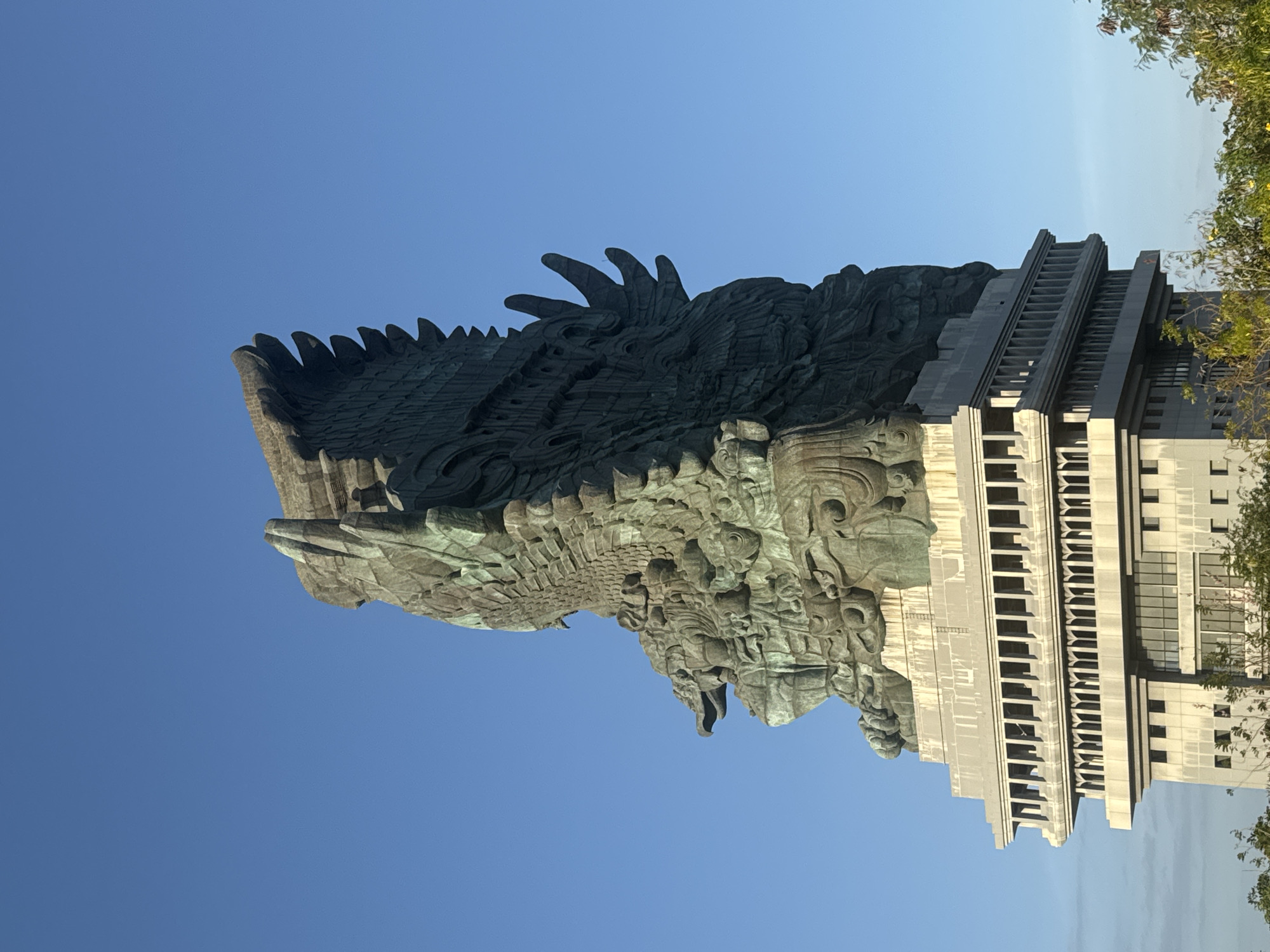 The image shows the Garuda Wisnu Kencana (GWK) Cultural Park in Ungasan, Kuta Selatan, Bali, Indonesia. The dominant subject is a colossal bronze statue of Vishnu riding Garuda, reaching high into a clear blue sky. The statue is intricately detailed, depicting Garuda's feathered wings, a reptilian head with a distinctive beak, and Vishnu's form with multiple arms. The statue is mounted on a multi-tiered pedestal that appears to be made of stone or concrete, with a colonnaded structure visible. Below the pedestal, parts of a building or the base of the monument are visible, with windows and architectural details. In the foreground, the tops of green trees and foliage are present, suggesting an outdoor setting. The lighting indicates it is daytime, likely morning or afternoon, with the sun illuminating the scene from the side, casting shadows and highlighting the statue's texture.