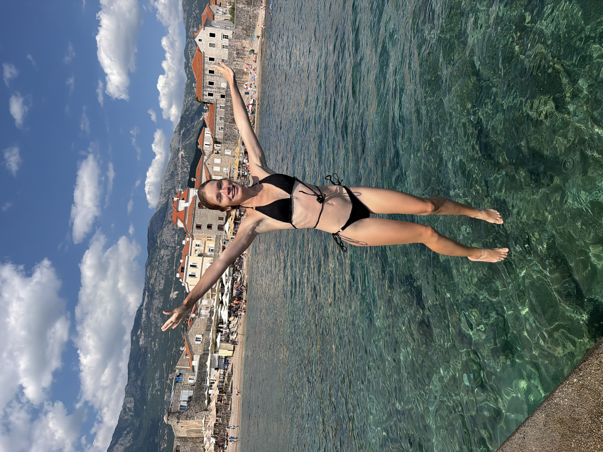 A young woman in a black bikini stands with her arms outstretched in the clear, shallow waters of the Adriatic Sea in Budva, Montenegro. The water is so clear that the rocky seabed is visible beneath the surface. In the background, the ancient walled city of Budva, with its terracotta-roofed buildings and stone walls, rises up from the coastline. Mountains form a dramatic backdrop to the scene, their slopes covered in green vegetation. A beach with umbrellas and sunbathers is visible to the left of the city. The sky above is a vibrant blue, dotted with fluffy white clouds, suggesting a warm, sunny day.