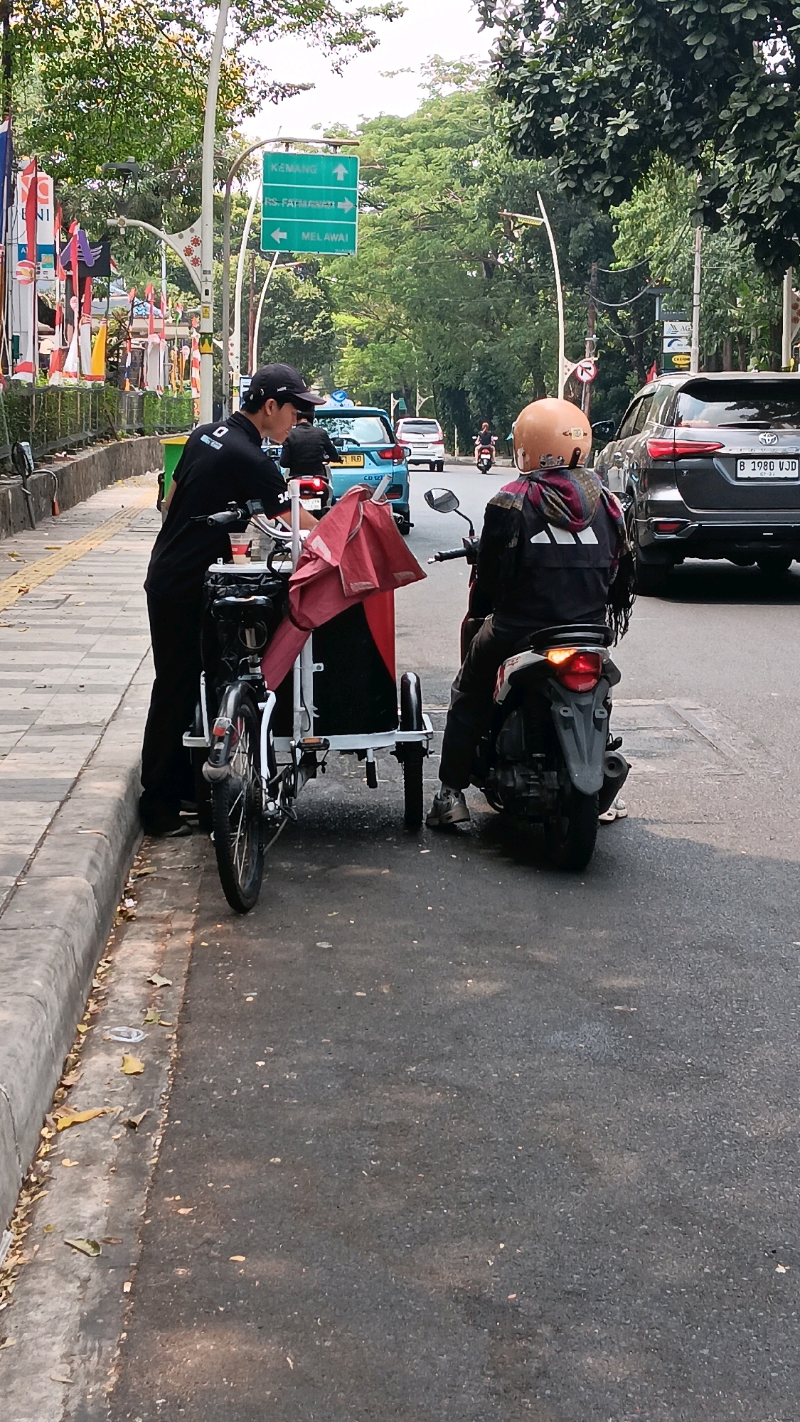 In the city of South Jakarta, Indonesia, a street scene captures two individuals on motorbikes by a sidewalk and roadway. A man wearing a black t-shirt and a black baseball cap is standing next to a three-wheeled bicycle, which has a red and black cargo box. He appears to be interacting with the bicycle. In the background, a person on a motorbike wearing a helmet and a black jacket with an Adidas logo is also present, facing away from the camera. Traffic signs indicating directions to Kemang, RS Fatmawati, and Melawai are visible above the roadway. Several vehicles, including a blue car with "CD 07" on its license plate, a dark SUV with license plate "B 1980 VJD", and other motorbikes, are present on the road. Trees and foliage line the street, suggesting an urban environment.