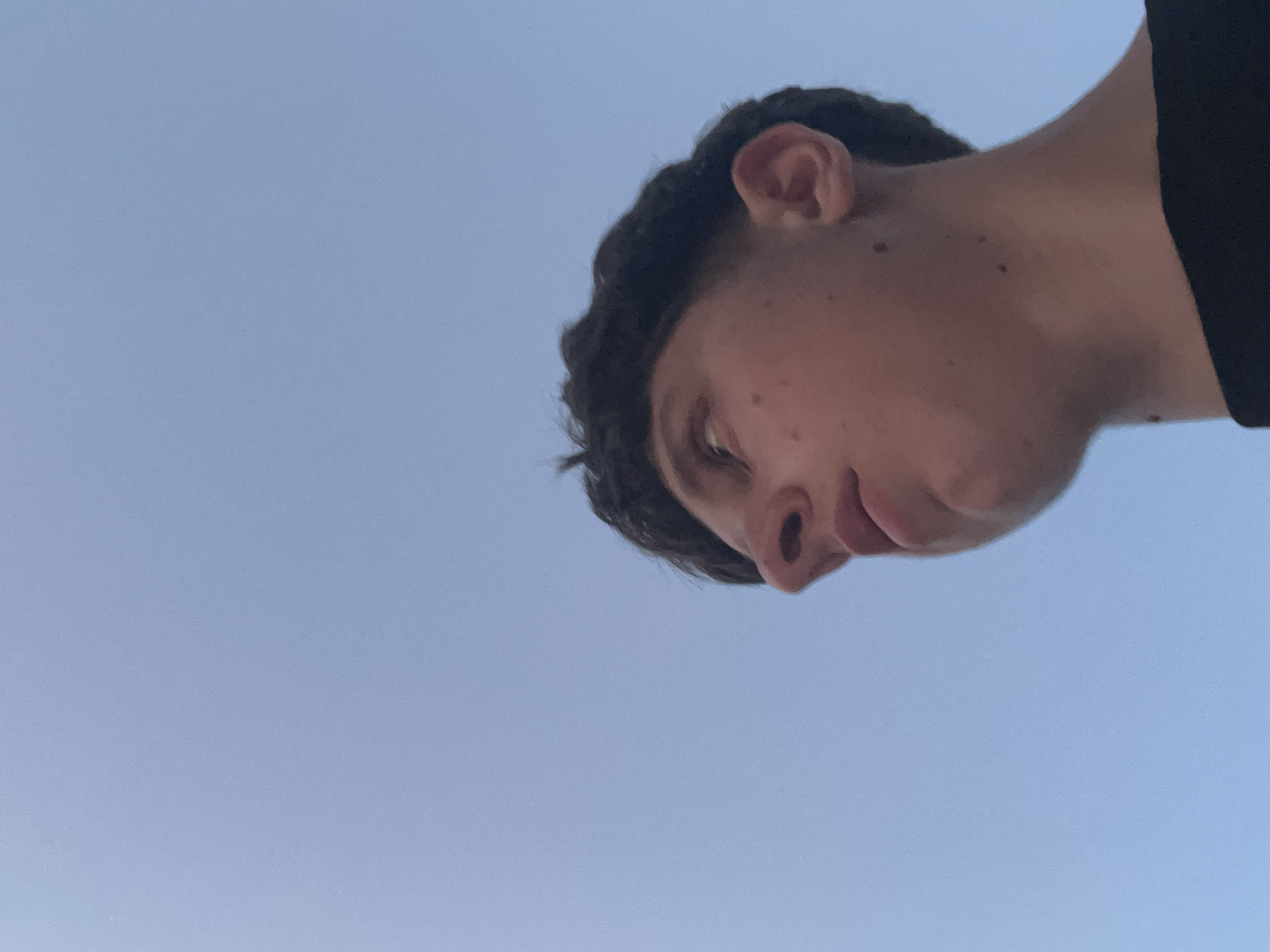 A low-angle, head-and-shoulders shot captures a young male against a clear, light blue sky in Zlatograd, Bulgaria. The subject, positioned in the lower right portion of the frame, has dark, wavy hair with a faded cut on the sides and back. His head is turned to his left (viewer's right) and tilted upwards, with his gaze directed towards the upper left. Visible features include a prominent nose, slightly parted lips, chin, and his right ear. Several small, dark moles are present on his right cheek and neck. He is wearing a dark, likely black, crew-neck t-shirt, the collar of which is visible at the bottom of the image. The background consists entirely of an unblemished, light blue sky, indicating an outdoor setting with clear weather. Natural light illuminates the subject from above and slightly to the right, creating subtle shadows.
