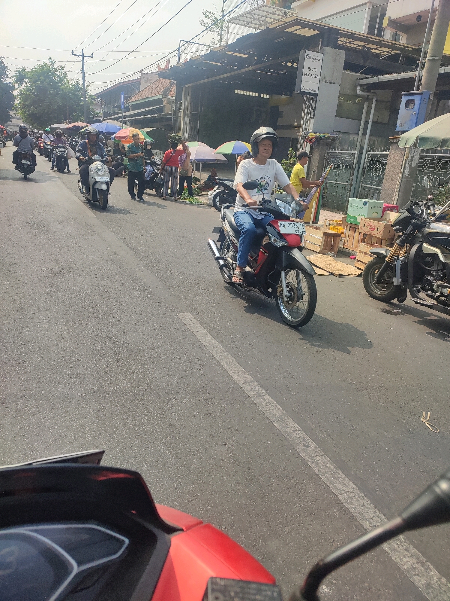In Yogyakarta, Indonesia, a street scene unfolds under a bright sky. A person on a red and black motorcycle, wearing a white t-shirt and blue jeans, is captured in motion. They are wearing a white helmet and appear to be looking forward. Behind this motorcycle, other vehicles, including motorcycles and scooters, are visible on the asphalt road, some carrying passengers. The roadside features buildings with awnings, suggesting a commercial area, and a sign that reads "ROTI JAKARTA". Several people are present, some standing and others on motorcycles, with umbrellas and market stalls indicating a lively environment, possibly a market or a busy thoroughfare. The overall impression is that of a typical day on a busy street in Yogyakarta.