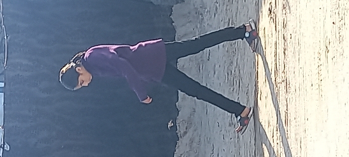 A person wearing a dark purple shirt and dark pants stands on a rough, light-colored surface. The person is leaning forward slightly. A dark, shadowy area is visible to the left, possibly a wall or building. The person wears dark shoes with red accents, and a watch is visible on one wrist. The surface appears to be textured and sunlit. The image is slightly rotated.