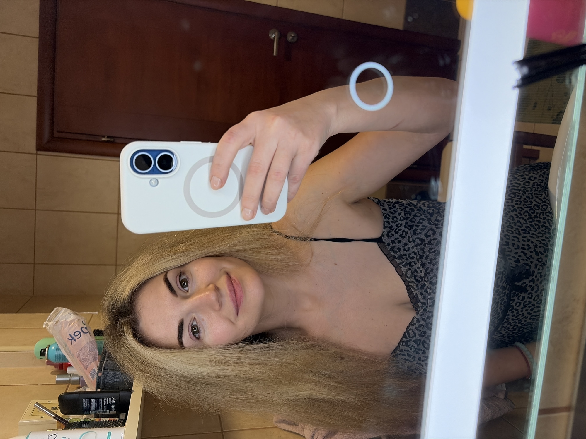 A young adult female is depicted taking a mirror selfie inside a bathroom, presumably located within a private residence or accommodation in Budva, Montenegro. The female has long, light blonde hair appearing slightly disheveled and is wearing a dark, leopard print camisole with black lace trim. She holds a white smartphone with a blue-rimmed dual-camera module and a visible MagSafe-compatible ring in her right hand. Her reflection shows her with a subtle smile.
The bathroom environment features light beige tiled walls. A wooden shelf on the left side holds various personal care products, including a tall white bottle, a dark deodorant stick, and a clear plastic bag containing items labeled "90k" in reverse. A dark brown wooden door with a metallic handle and lock is visible in the reflection on the right side. Below the mirror, a horizontal white light strip illuminates the scene, with a glass shelf beneath it. A red bracelet and a light blue/green beaded bracelet are visible on the female's left wrist.