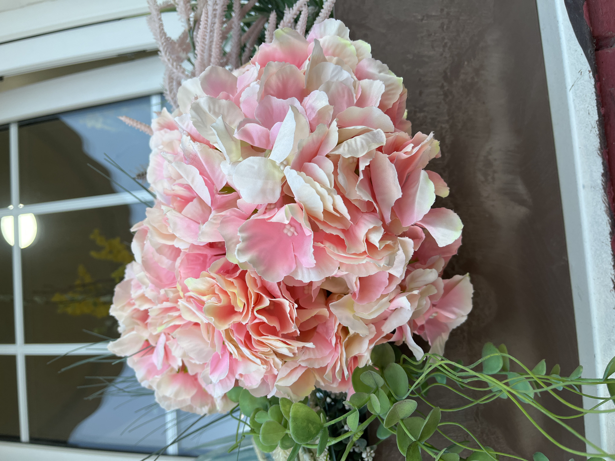 A close-up shows a large artificial floral arrangement featuring pink and white flowers resembling hydrangeas. Green leaves and sprigs are visible, adding depth. The arrangement is positioned against a brown-textured wall, with a portion of a white-framed window and brick visible on the left and right sides of the frame, respectively. The image does not provide information to confirm it was taken in Podolsk, Russia.