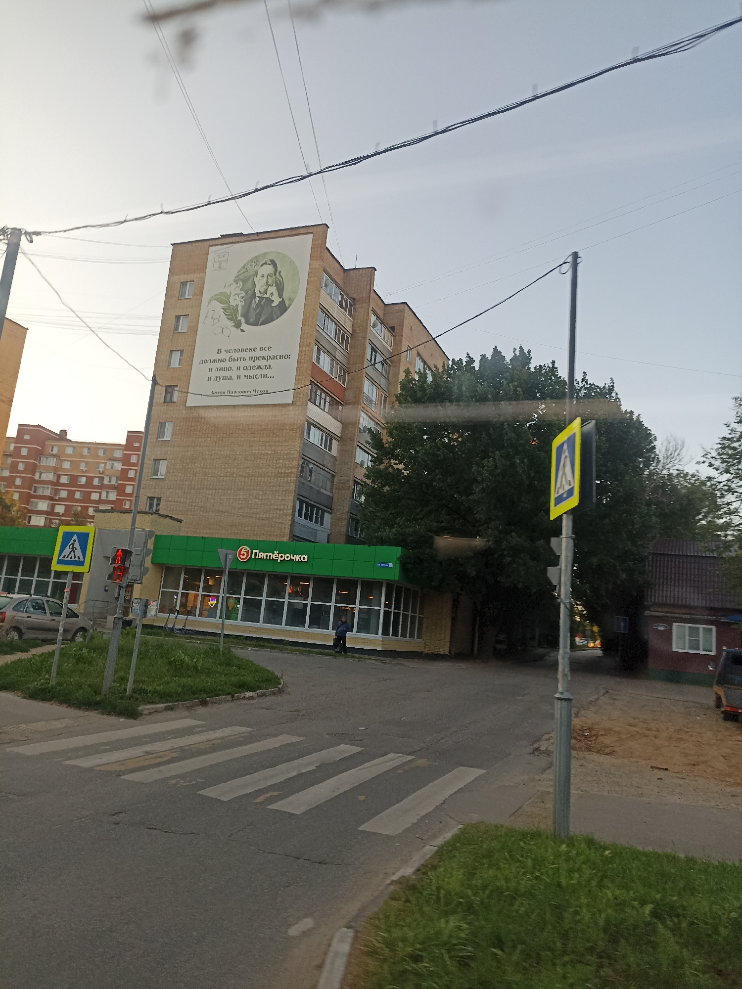 The image depicts a street scene in the city of Chekhov, Russia, with a focus on a tall brick building featuring a large mural. The mural is circular and displays a portrait of a man, accompanied by text that appears to be a quote. Below the mural, the building has multiple floors with visible windows. In front of the building, there is a "Pyaterochka" supermarket with a green awning and large windows. A pedestrian crossing is visible in the foreground, marked by white stripes on the asphalt. A traffic light is present, showing a red signal. A yellow pedestrian crossing sign with a blue symbol is mounted on a pole. Several cars are parked or driving on the street. Mature trees are growing behind the supermarket, and some smaller residential buildings are visible in the background. The sky is overcast, suggesting it may be late afternoon or early morning. A person is walking near the supermarket entrance.