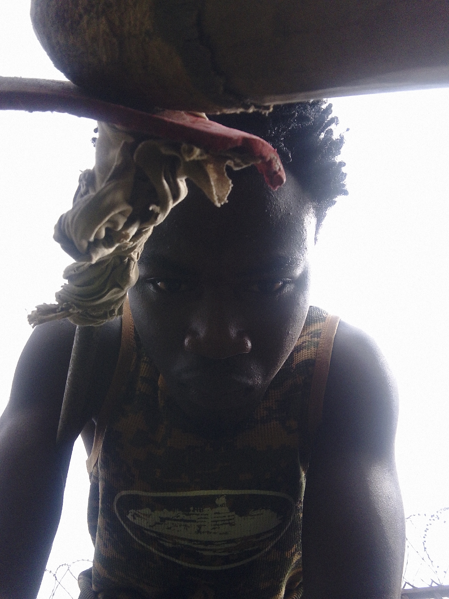 A low-angle, close-up shot depicts a person in Barikin Gidar Kanat, Nigeria. The individual, appearing to be male, has dark skin and short, textured hair. A light-colored cloth is draped over their head. They are wearing a camouflage-patterned tank top with a graphic design on the front. The background is bright, almost washed out, suggesting outdoor daylight. A cylindrical object and a red bar are partially visible at the top left of the frame. Faintly visible in the background is what appears to be a barbed wire fence.