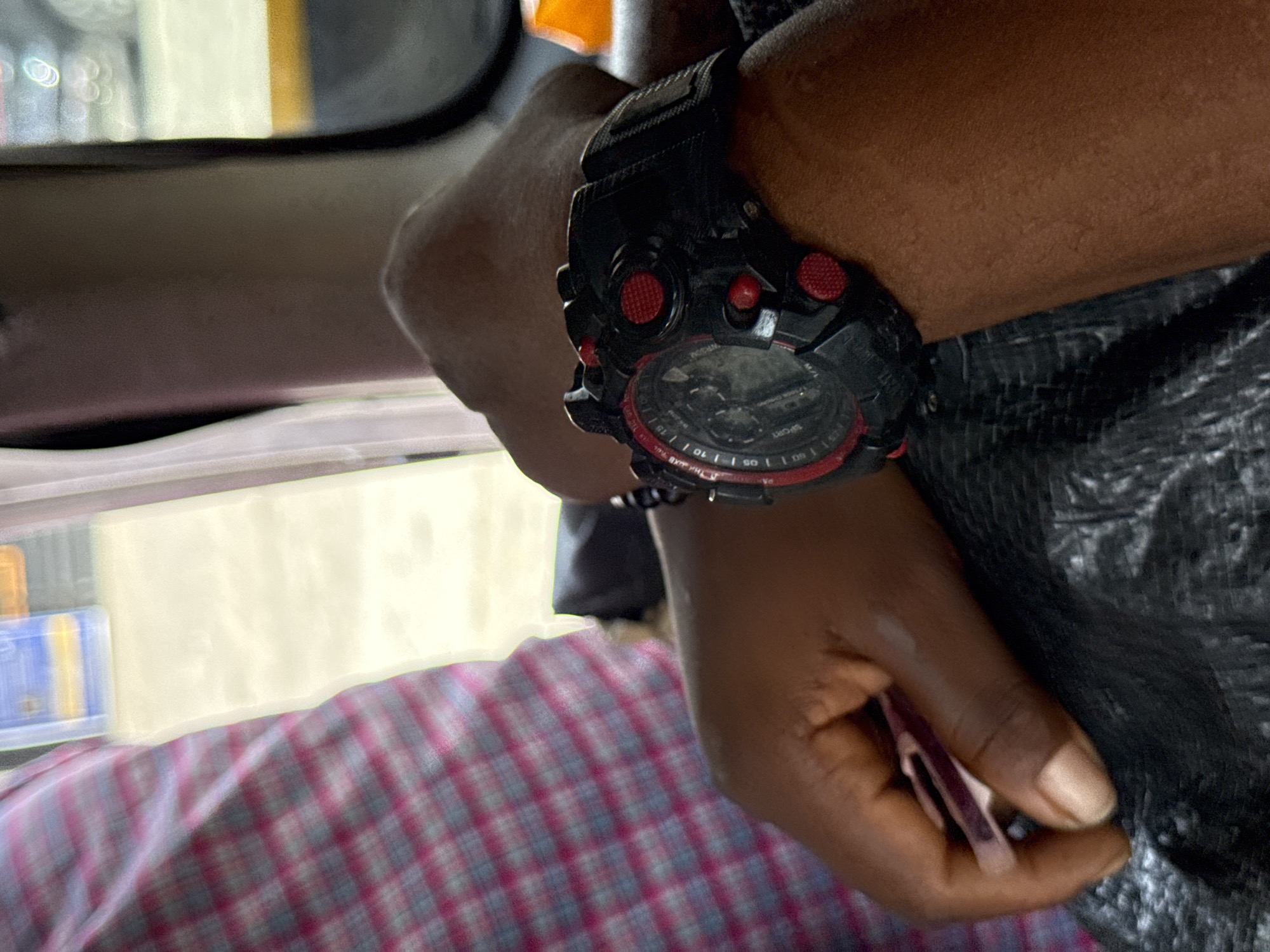 A close-up shot shows a person's arm wearing a large black digital watch with red accents. The person is holding a small, pink object in their right hand. The person is wearing a plaid shirt. The location appears to be inside a vehicle, possibly a car or taxi. The background is blurred, but suggests an urban environment, potentially Ilado, Nigeria.