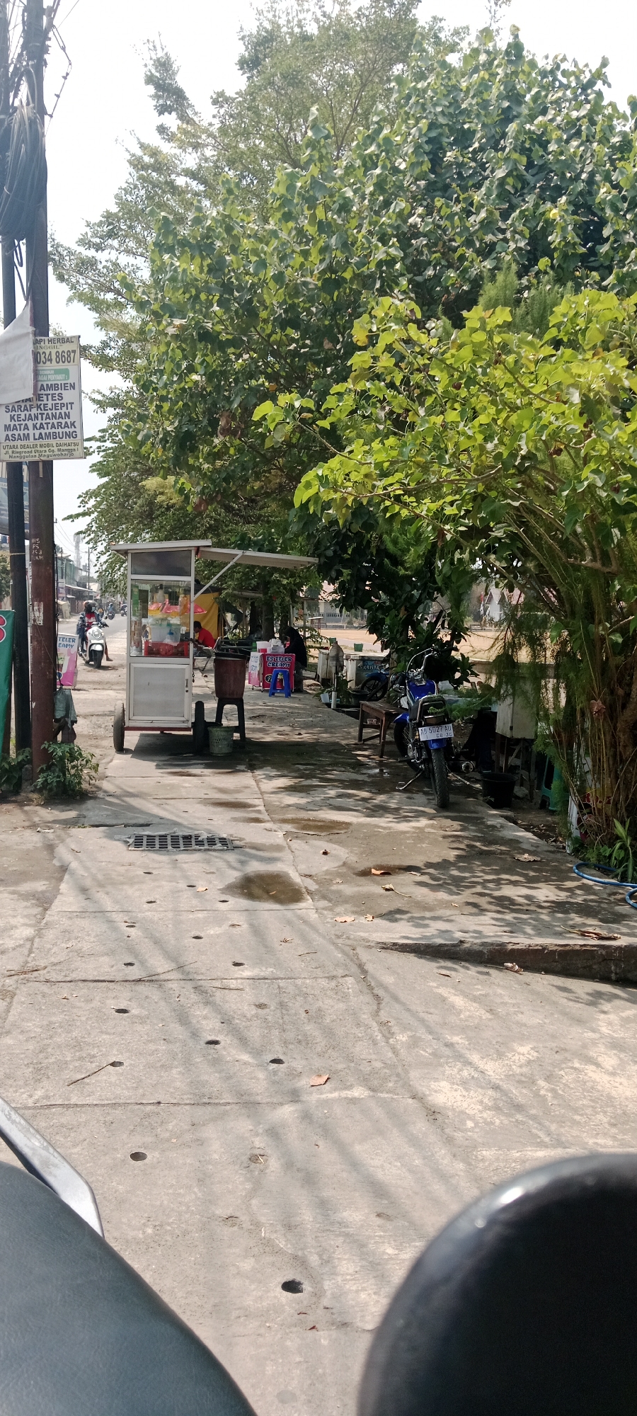 This is a vertical, medium-distance, outdoor shot of a street scene in Maguwoharjo, Indonesia, under bright daylight conditions. A concrete sidewalk runs diagonally from the foreground upwards and to the right, shaded in places by large, leafy trees. On the left side of the sidewalk, a vertical utility pole with cables is visible. To the right of the pole, a white and red food cart with a canopy is parked, displaying colorful drinks and food items inside a glass enclosure. A person in a red shirt is standing behind the cart. A motorcycle rider in a black helmet is seen on the street to the left of the cart, approaching the viewer. Further down the sidewalk, to the right of the cart, is a blue plastic stool next to a small table. Behind this, there are a few more people seated and standing. A blue motorcycle is parked on the right side of the scene, facing away from the viewer. The street itself is paved with concrete slabs, with a metal grate and some scattered leaves visible. The overall impression is of a typical street scene in a commercial or residential area.