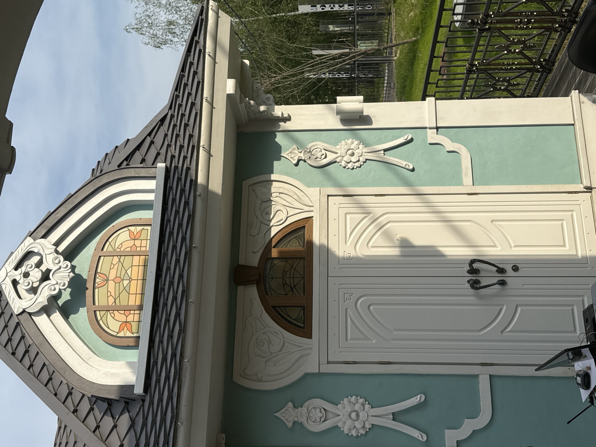 This is a close-up, eye-level outdoor photograph of the facade of a small, ornately decorated building with a mint green exterior wall. The building has a steeply pitched dark gray roof with diamond-shaped tiles and a white decorative trim along the eaves. A small, arched stained-glass window with a floral motif in shades of orange, yellow, and green is visible on the upper part of the facade, framed by white architectural embellishments. Below this, a large, white double door with carved panels and dark metal handles dominates the lower part of the image. To the left and right of the door, decorative white plaster moldings in a stylized floral and ribbon design adorn the wall.
A black metal fence is visible to the right of the building, with a grassy area and trees beyond. In the foreground, on the left side of the image, a small table or counter holds a laptop, a radio transmitter, and other electronic devices.
This building is located in Moscow, Russia. Specifically, it is part of the Serebryany Bor recreational area, a protected natural reserve located on an island in the Moskva River in the west of the city. This particular structure is a small, decorative pavilion or kiosk within the park.