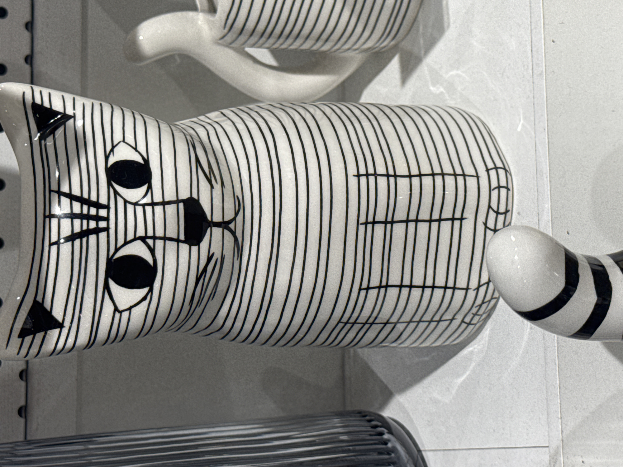 A close-up shot focuses on a black and white striped ceramic cat figurine, positioned on a white surface that appears to be a shelf or countertop. The figurine's face features large, round eyes with black pupils, triangular ears, and subtle lines suggesting a nose and whiskers. Its body is covered in thin, horizontal black stripes, with vertical lines indicating legs. A separate, detached piece, likely a tail, with similar black and white stripes, lies in the foreground. To the left of the cat figurine, a tall, fluted glass vase with vertical ridges is partially visible, casting a shadow on the surface. In the background, to the right of the cat, another white ceramic object, possibly a mug or pitcher, also adorned with black stripes, is visible. The environment suggests an indoor setting, possibly a retail display or a home. The image is devoid of people or any indication of activity. There is no information available to determine the location in the city of Ufa, Russia.