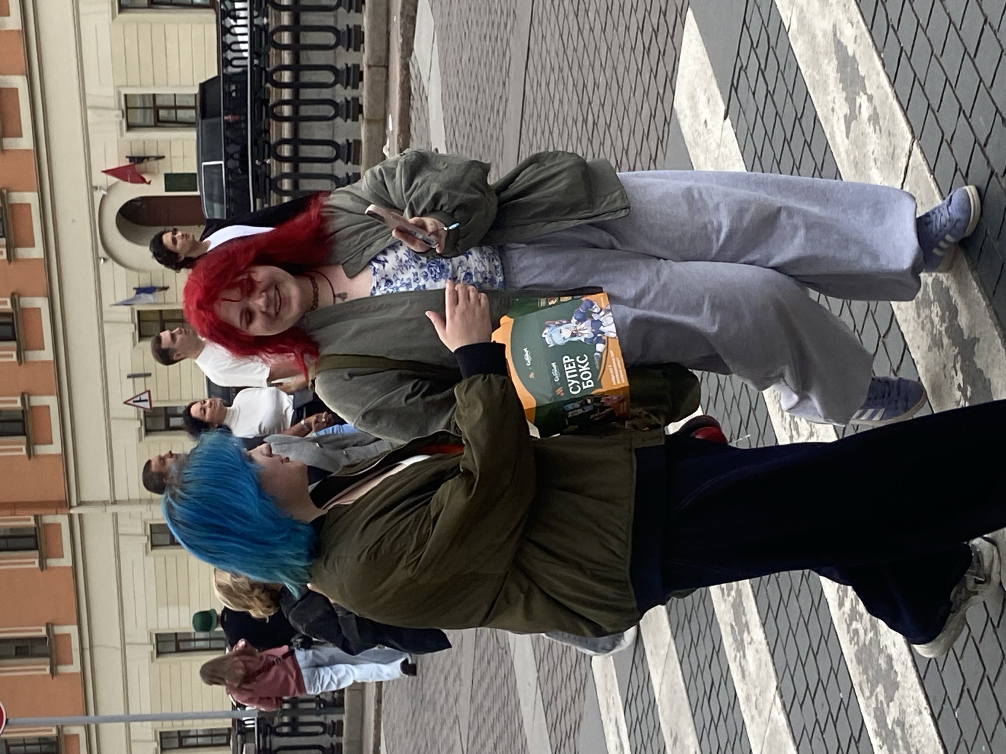 Two young women are crossing a street in Saint Petersburg, Russia. The woman on the left has bright blue hair and is wearing a dark green bomber jacket over a white top and dark pants. The woman on the right has vibrant red hair and is wearing a light blue floral-patterned top, a light olive green bomber jacket, and light grey sweatpants. She is holding a smartphone in her right hand and carrying a brightly colored box, possibly a gift or promotional item, with Russian text that reads "СУПЕР БОКС" (Super Box) and an image of a cartoon character. They are walking on a pedestrian crossing with black and white stripes. In the background, other pedestrians are visible, along with a building with an orange facade and arched windows, and a black car parked behind a decorative metal fence. The overall scene suggests a casual urban environment.
