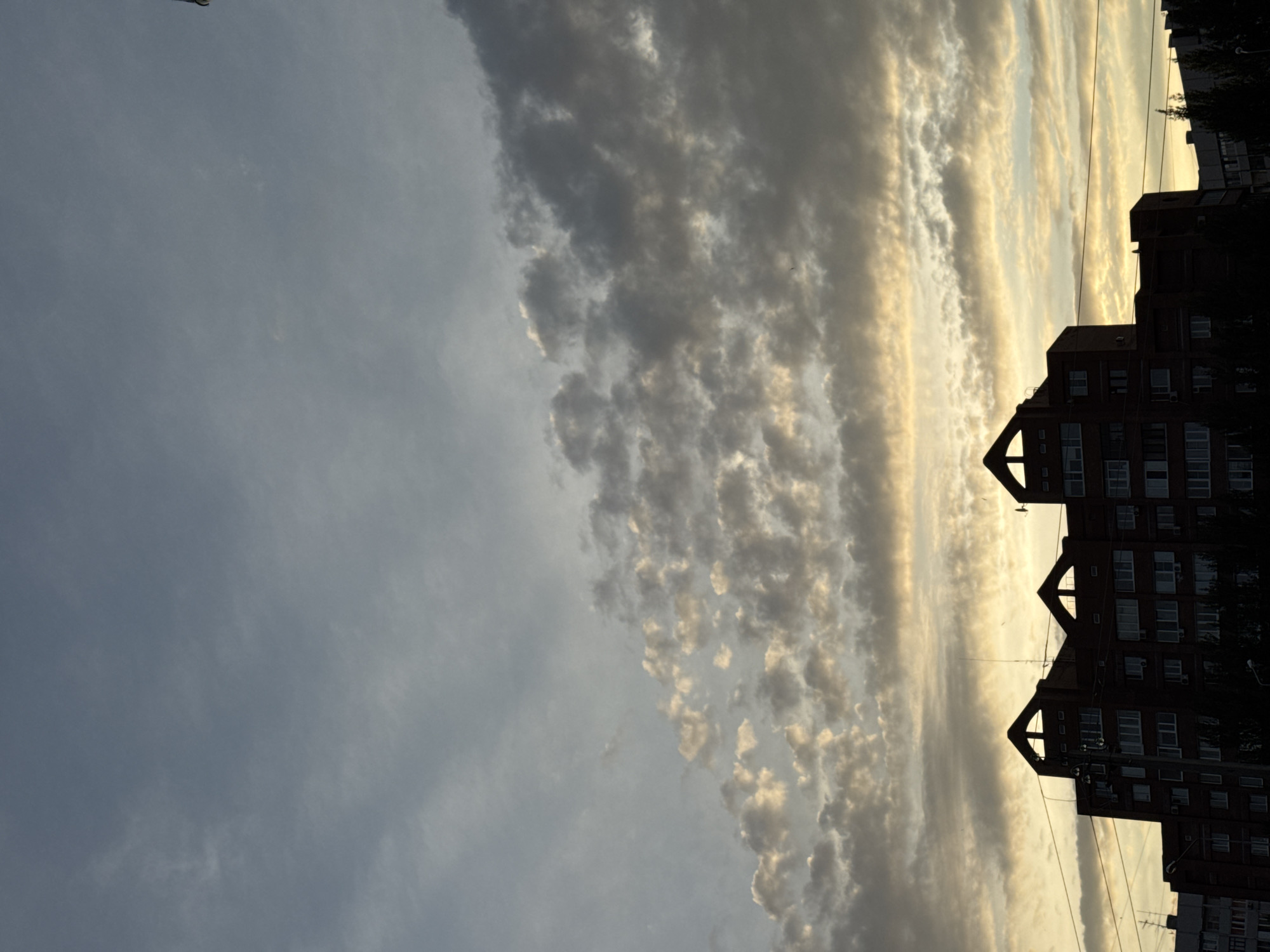 The image captures a silhouette of apartment buildings against a cloudy sky. The buildings are dark, with visible windows and distinct architectural features on their rooftops. The sky is a blend of grays and yellows, suggesting either dawn or dusk. There are no people or overt actions depicted, focusing solely on the urban landscape and atmospheric conditions. The scene is static and devoid of any specific contextual markers that would pinpoint a precise location within a city.