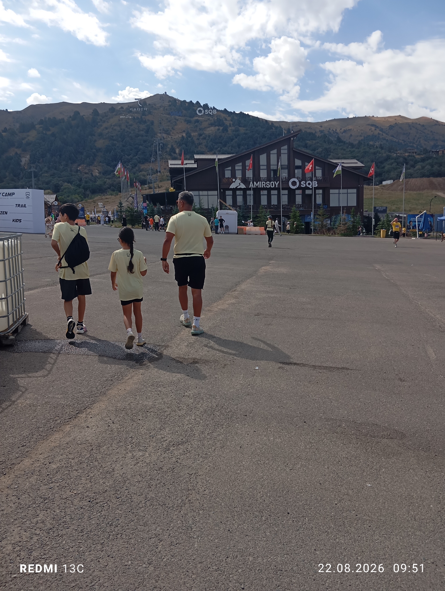 In the city of Amirsay, Uzbekistan, which is a ski resort, three people are walking away from the camera across an asphalt-paved area. The tallest individual on the right, a man, is wearing a light yellow t-shirt and black shorts. To his left, a young girl with dark hair in a ponytail, also wearing a light yellow t-shirt and black shorts, walks slightly ahead. On the far left, a young boy, wearing a similar light yellow t-shirt and dark shorts, carries a black backpack. In the background, a large, modern building with a dark brown exterior and large windows displays the text "AMIRSOY SQB" above its entrance. Mountains covered in trees form the backdrop, with a clear blue sky dotted with white clouds overhead. Several flags are visible in front of the building, and other individuals are scattered throughout the scene, some appearing to be walking or running. A large white container is situated to the left of the frame. The timestamp in the bottom right corner indicates "22.08.2026 09:51".