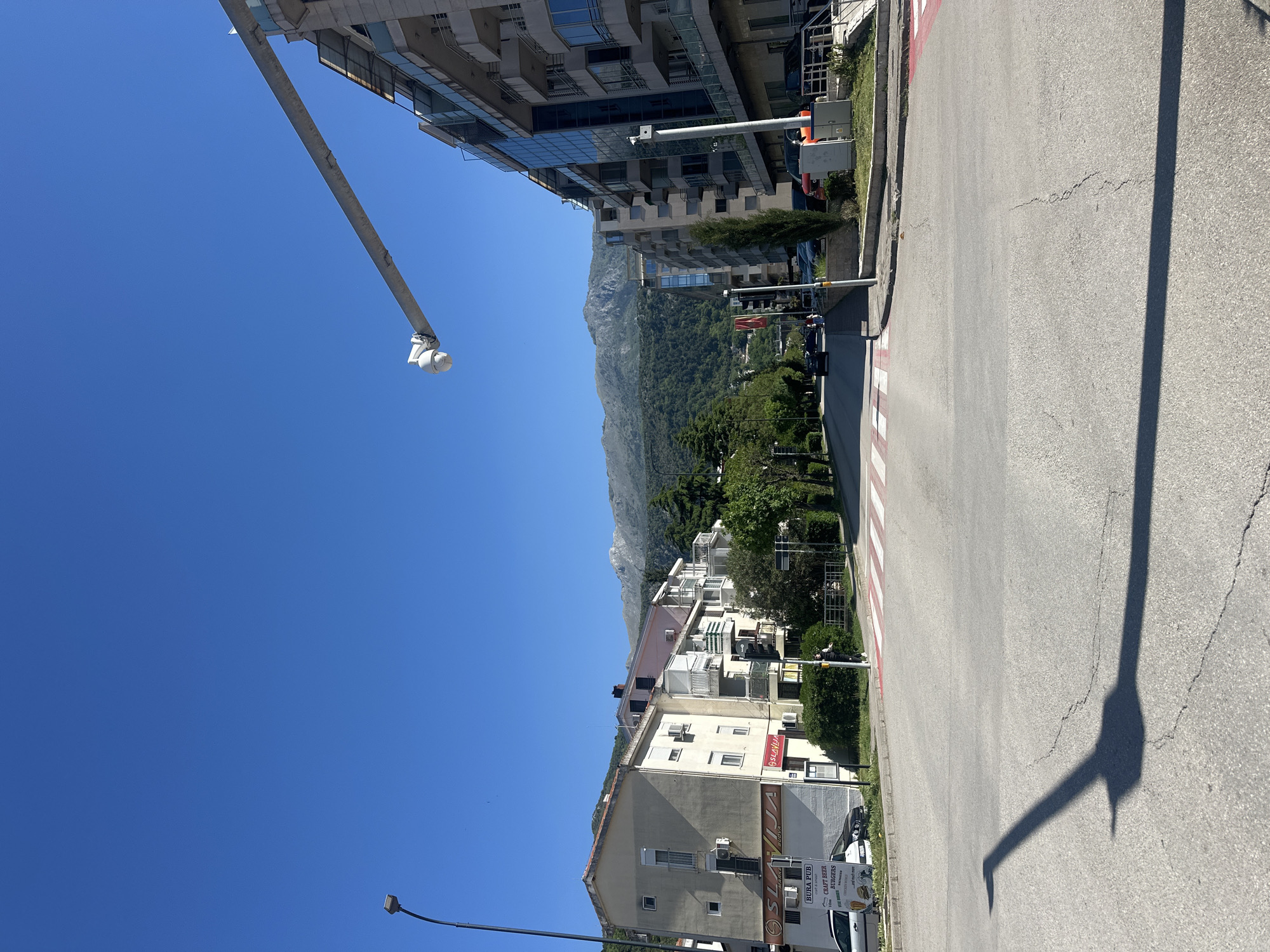 The image captures a bright, sunny daytime scene in what appears to be an urban area, with mountains in the background, consistent with the provided location of Budva, Montenegro. The sky is clear blue with no clouds.
The perspective is from a street level, looking towards a mountainous landscape. On the left side of the road, there is a modern, multi-story building with a glass facade and numerous balconies. Extending from this side, a long arm with what appears to be a camera or sensor points outwards over the road. On the right side of the road, there are older, residential-style buildings, also featuring balconies and visible air conditioning units. The mountains in the background are covered in dense green vegetation.
The asphalt road features a distinct pedestrian crossing with red and white stripes. A prominent, elongated shadow of a streetlight pole is cast across the road from the right side, indicating the sun is high in the sky. Utility boxes and some greenery are visible along the roadside.
Visible text on a sign on one of the buildings on the right includes "BURA PUB," "CRAFT BEER," and "BURGERS," along with "cycle & lounge" and "restaurant." Another sign above it partially reads "SSLLAVIJA" or "SLAVIJA" and "BUONA" or "NUOVA." No explicit human activity is captured, but the scene depicts a typical street environment in a town or city.