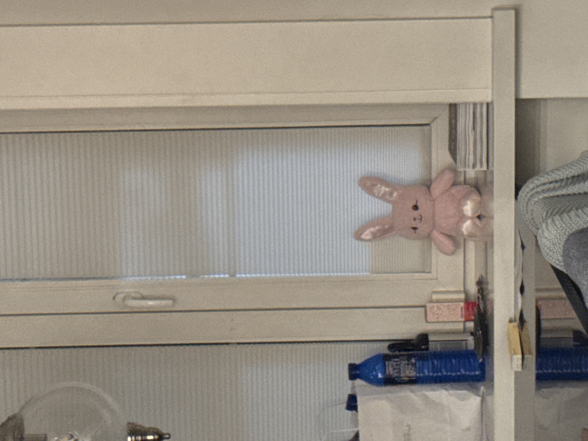 A stuffed pink bunny with a bow sits on a windowsill in front of a closed window with horizontal blinds. To the left of the bunny, a blue bottle and a white paper bag are visible on the windowsill. A stack of books or papers is to the right of the bunny. The windowsill is above a shelf where a light blue knitted item is draped.