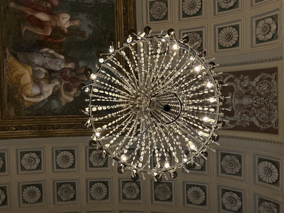 The image displays an upward view of a richly decorated ceiling within a grand interior, identified as the Palazzo Clerici in Milan, Italy.
A large, multi-tiered crystal chandelier, centrally positioned and illuminated, dominates the lower half of the frame. It features numerous clear, faceted crystal beads and pendants strung between intricate metal frames, with multiple incandescent, candle-style bulbs emitting a warm light.
In the upper right and center, a rectangular ceiling fresco is visible, bordered by an ornate gilded frame. The fresco depicts a mythological or allegorical scene with multiple draped figures in classical poses, some reclining or interacting, set against a background of foliage and sky. The figures are rendered with a palette of blues, greens, yellows, and reds.
To the left and bottom of the fresco, extending across the upper and lower left of the image, is a section of a coffered ceiling. This ceiling consists of regularly spaced square panels, each framed by decorative molding and containing a raised, pale relief of a floral rosette or medallion. The background of these panels is a light greenish-grey.
Directly below the fresco and partially visible in the lower center, an elaborate section of bas-relief stucco decoration is present. This features intricate white scrollwork, acanthus leaves, and classical motifs, possibly including a central urn or vase, set against a reddish-brown painted background. The entire scene indicates an opulent, historic European palace interior.