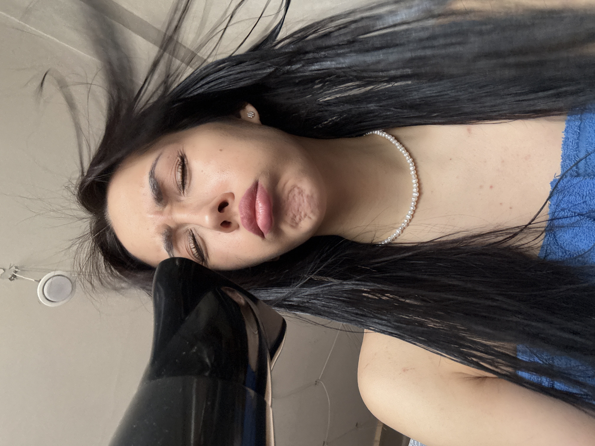 A woman with long black hair is drying her face with a black hairdryer. She is wearing a pearl necklace and a blue towel. Her eyes are closed and her face is scrunched up. The background is a light beige wall.
I cannot provide the location in the city of Votkinsk, Russia, as the image does not contain that information.