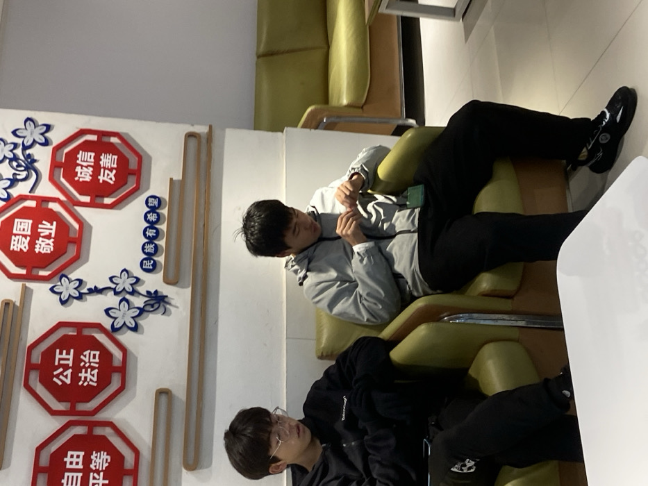Two people lounge in adjacent green chairs. One person wears a grey jacket, black pants, and black shoes. They recline with their legs stretched out. The second person wears a black hoodie and black pants. On the wall are several red octagonal shapes with Chinese characters and floral decorations.