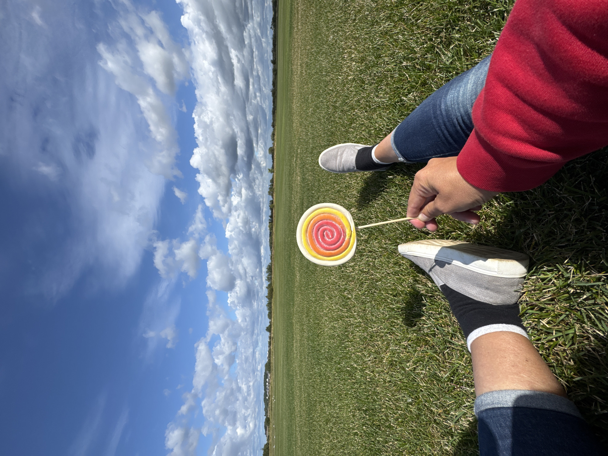 The image shows a person sitting on a green grassy field under a blue sky with scattered clouds. The person is holding a colorful lollipop with red, orange, and yellow swirls. Their legs are visible, one wearing a red long-sleeved shirt, blue jeans, and black socks with white trim, and the other leg wearing a grey slip-on shoe and black socks. The field stretches out to the horizon, with a line of trees and some distant buildings. The scene is calm and suggests an outdoor recreational activity. The location is identified as Innolovo, Russia.