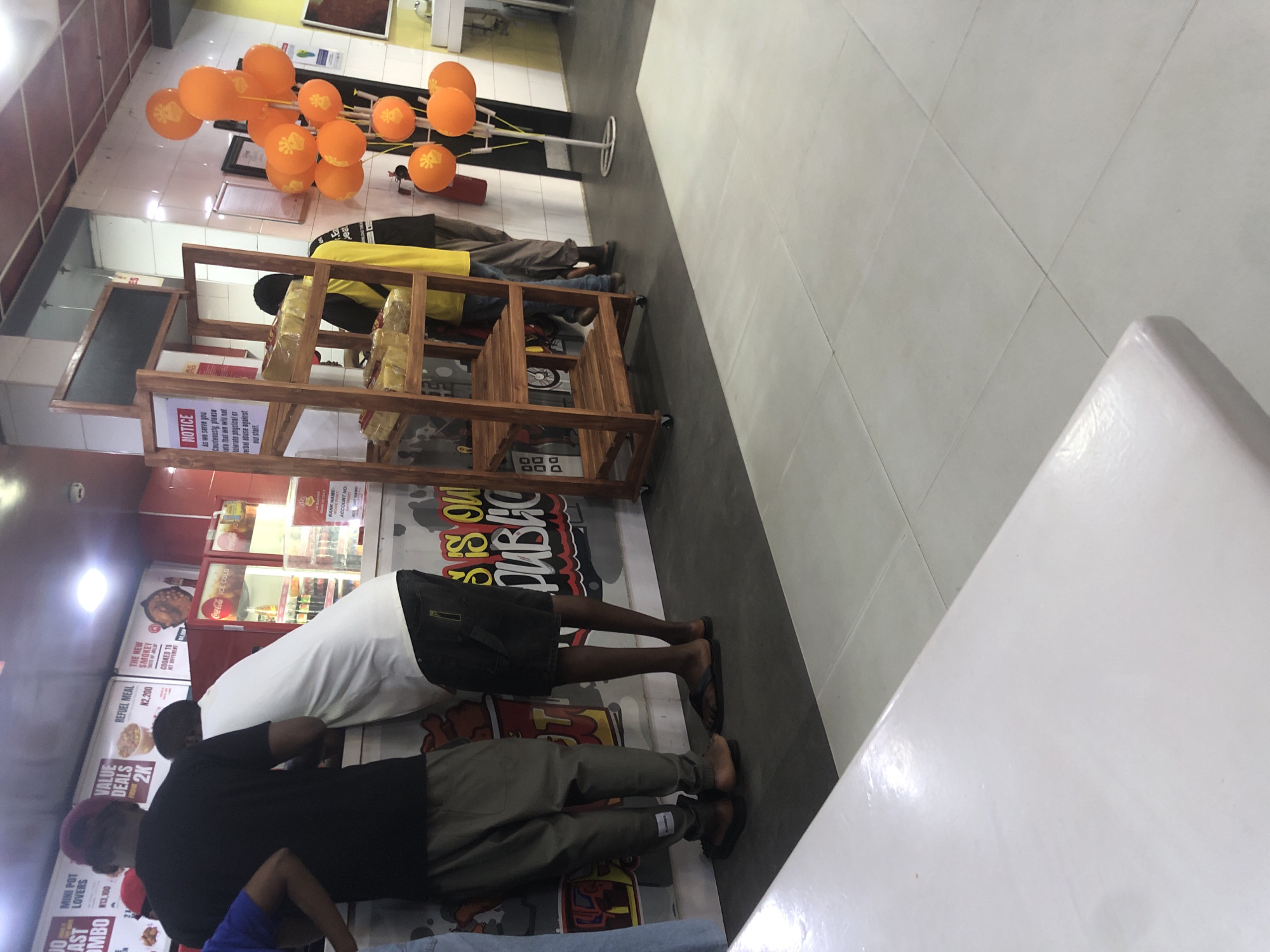 The image depicts a fast-food restaurant, likely in Ado, Nigeria, based on menu items and prices visible. A group of people, predominantly male, are gathered at the counter, appearing to order or wait for food. One individual, wearing a dark t-shirt and a maroon cap, is seen from behind. Next to him, another person in a white t-shirt and dark shorts stands by the counter. Behind them, a person in a yellow shirt and dark pants is partially visible, looking towards the counter.
Behind the counter, a refrigerator displaying "Coca-Cola" branding is visible, stocked with drinks. A wooden display rack with several food items, possibly bread or pastries, is positioned to the right of the counter. A "NOTICE" sign is attached to the rack, providing instructions to customers.
To the right of the rack, a cluster of orange balloons with an emblem is displayed on a stand. The background features walls with colorful graphics, including text that appears to say "SIS OUT PUBACO" and other unintelligible words. The flooring consists of dark grey tiles near the counter area, transitioning to lighter grey tiled floors. The overall lighting suggests an indoor environment, possibly illuminated by fluorescent lights.