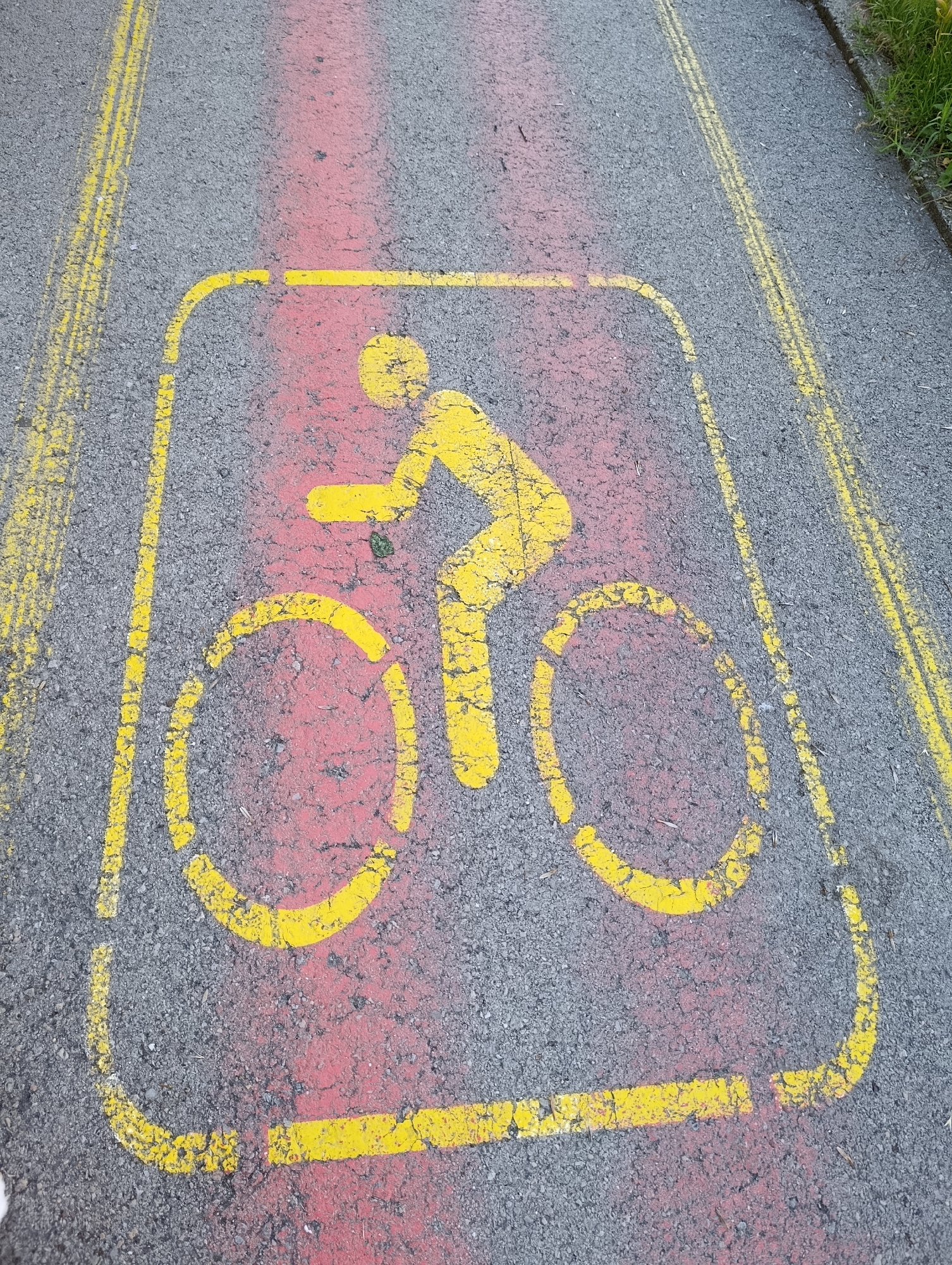 The image shows a yellow bicycle symbol painted on asphalt in Razgrad, Bulgaria. The symbol is enclosed in a yellow rectangle and consists of a cyclist figure riding a bicycle. Red paint has been applied underneath the yellow symbol. Two parallel yellow lines, likely lane markers, are visible on the left and right sides of the image. The asphalt surface appears worn and cracked in places. Green grass and a curb are visible along the right edge of the image, indicating the presence of a roadside or pathway. The lighting suggests it is daytime.