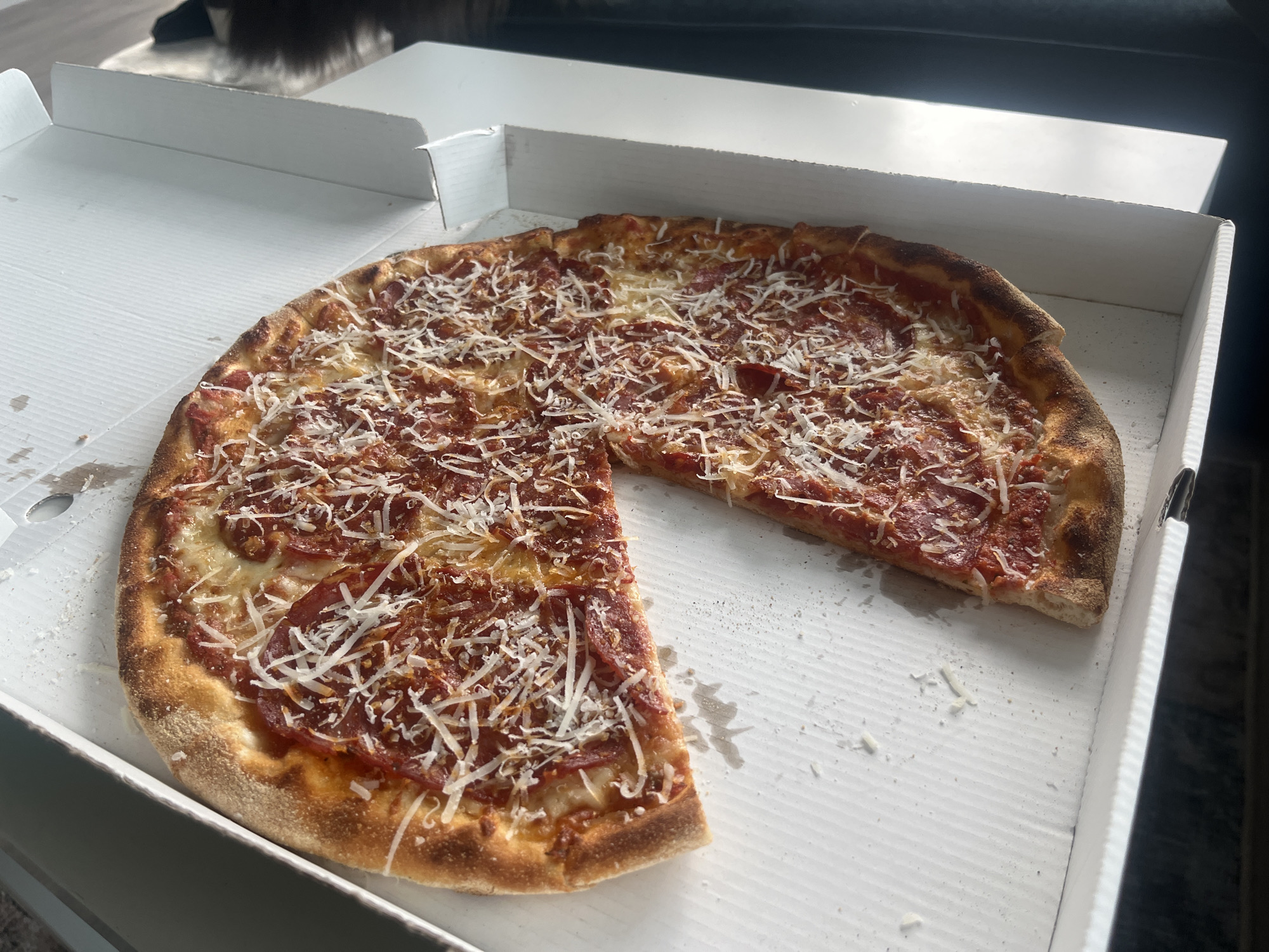 A high-angle forensic photograph displays an approximately 14-inch, partially consumed pizza situated within an open white corrugated cardboard pizza box. The pizza features a golden-brown crust, a red tomato sauce base, melted white cheese, numerous slices of red pepperoni or salami, and a liberal application of grated white cheese, likely Parmesan, dusted across its surface. One slice of the pizza has been removed, creating a triangular void. Scattered cheese crumbs and minor grease stains are visible on the white interior of the pizza box, particularly in the area where the slice is missing. The pizza box rests on a light-colored, possibly wooden, surface. In the blurred background, a dark piece of furniture or upholstery is visible on the right, and a light-colored floor or carpet is partially discernible on the left. The scene is illuminated by ambient indoor light. No individuals are present in the image. The location is in the city of Unknown, Unknown.