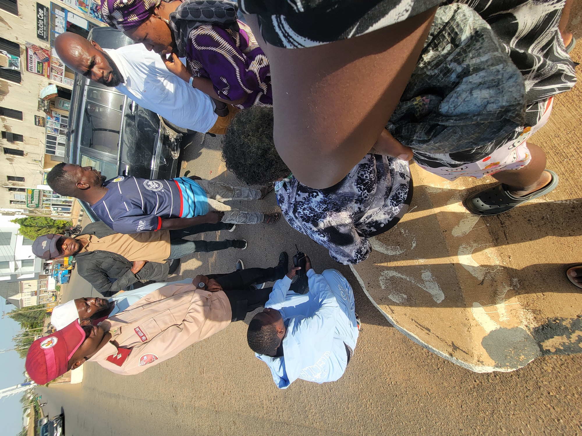 The image shows a group of people standing on a paved street in Barakin Lerawi, Nigeria. Several individuals are gathered together in a close configuration with at least seven distinct people visible from above in the photograph. Some individuals are wearing traditional Nigerian clothing such as a purple and white head wrap, a red cap and a light pink uniform with red accents and a white cap. One person is wearing a blue soccer jersey. An older-model black Mercedes car is parked nearby. Behind the car, a building has various store signs, including one for "Double Cee" cosmetics and "ROL FARMS" customer outlet. Additional buildings are also visible in the background. The environment suggests a commercial area within the city.