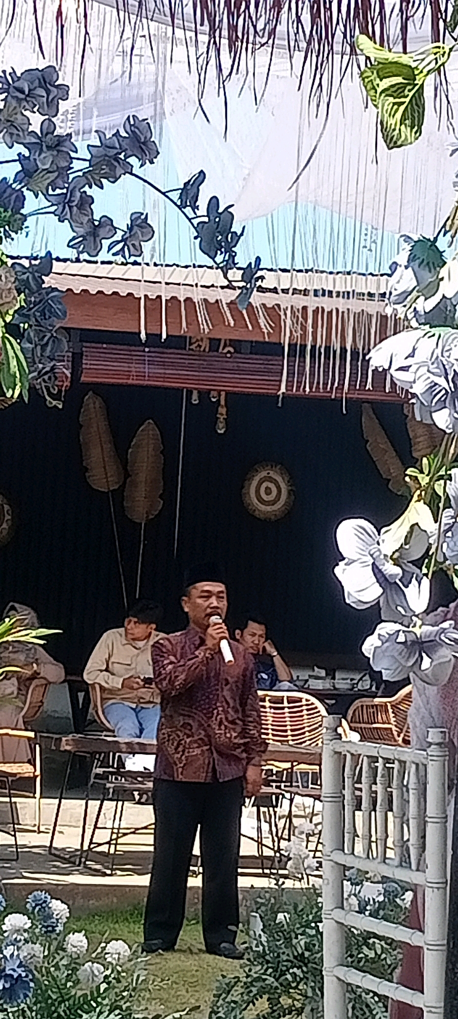 This image depicts an outdoor scene in what appears to be a cafe or event space in Desa Ponokawan, Indonesia. A man wearing a black peci (cap) and a patterned maroon shirt is speaking into a microphone while standing in the foreground. He is wearing dark pants and dark shoes. Behind him, several other individuals are seated at tables. One man in a light-colored shirt and blue jeans is looking at his phone. Another man with short dark hair is visible further in the background. The setting features a dark-colored wall adorned with decorative items, including what seem to be woven palm fronds and circular wall hangings. Overhead, a thatched roof or canopy is visible, with decorative fringe hanging down. Lush greenery, including flowers and leaves, frames the scene in the foreground and to the sides, suggesting an outdoor garden or landscaped area. White bamboo-like fencing is present on the right side of the image. The overall atmosphere appears to be relaxed, possibly during a speech or performance in a pleasant outdoor environment.