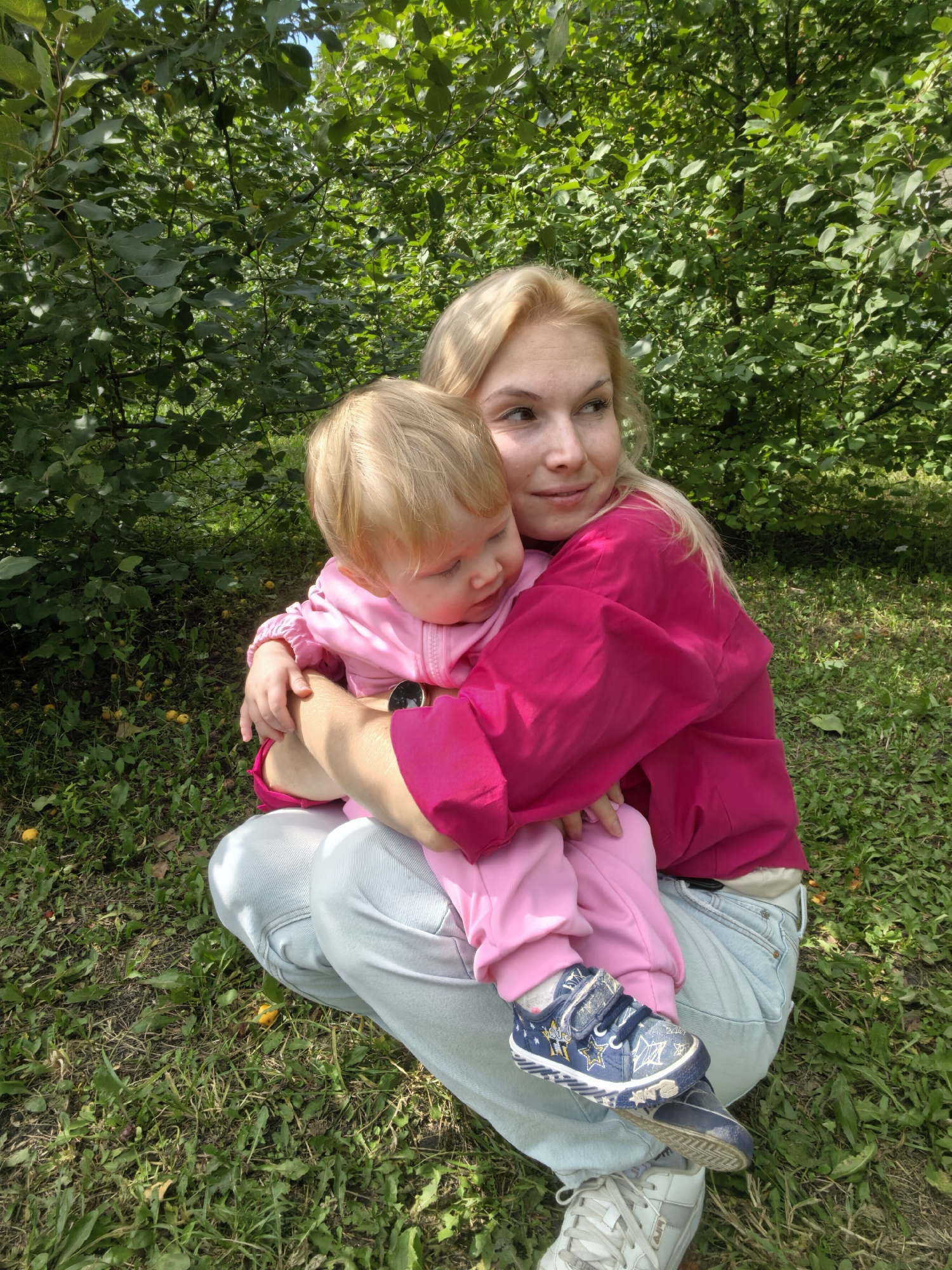 In Sterlitamak, Russia, a blonde woman kneels on the grass, embracing a young child also with blonde hair, who is resting their head on the woman's shoulder. The woman wears a bright pink top and light blue jeans, while the child is dressed in pink clothing and blue sneakers with star patterns. They are surrounded by lush green foliage and small yellow fruits scattered on the ground. The scene appears to be set in a park or garden on a sunny day.