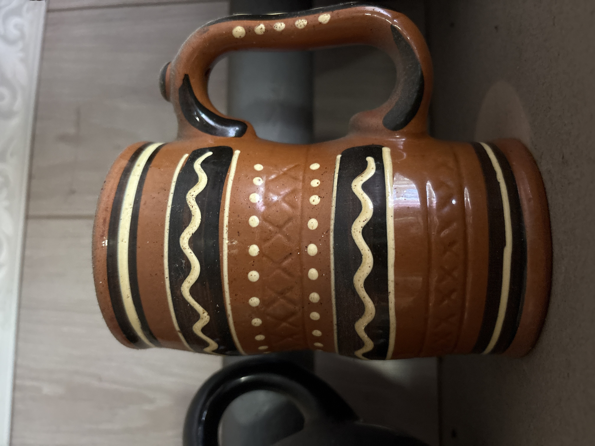 A close-up shot captures a brown ceramic mug with black and cream-colored decorative bands. The mug is cylindrical with a sturdy handle. The decorations consist of horizontal lines in black and cream, with a wavy cream pattern on a black band and a textured pattern with small cream dots in between. A portion of a black object, possibly another mug or container, is visible to the left of the brown mug. The objects are placed on a shelf against a backdrop of vertical wooden planks, suggesting an indoor setting. The lighting is somewhat dim, with highlights reflecting off the ceramic surface. The location is identified as Ветлужский, Russia.