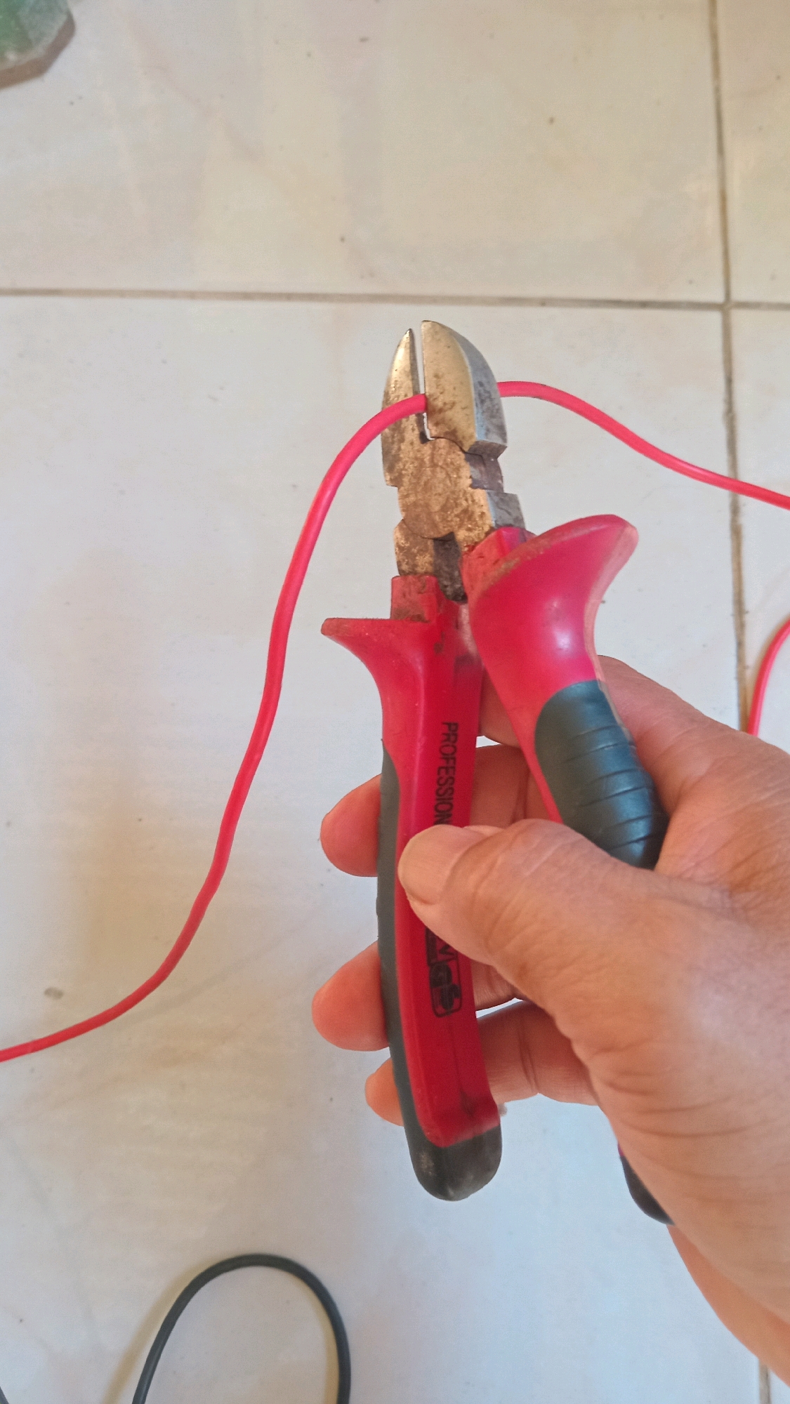 A person's hand is holding a pair of red and gray pliers. The pliers are positioned on a tiled surface and are cutting a red wire. The wire is looped and extends out of frame. The background is a light-colored tiled floor. The scene appears to be in an outdoor or semi-outdoor area given the lighting.