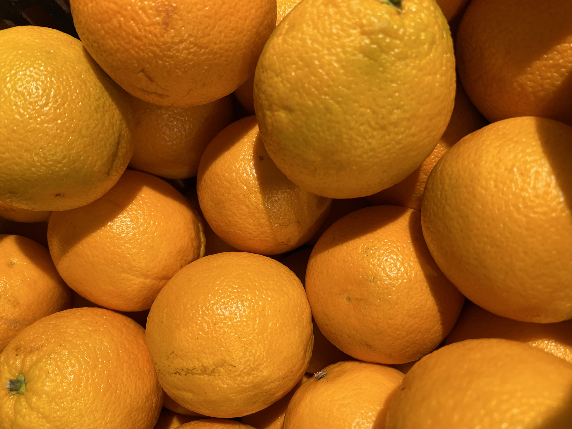A close-up, high-angle shot shows a pile of ripe oranges. The oranges fill the frame, creating a dense, textured pattern of round shapes and orange hues. The lighting suggests an indoor setting, possibly a market or grocery store. The scene contains no people or any other objects.