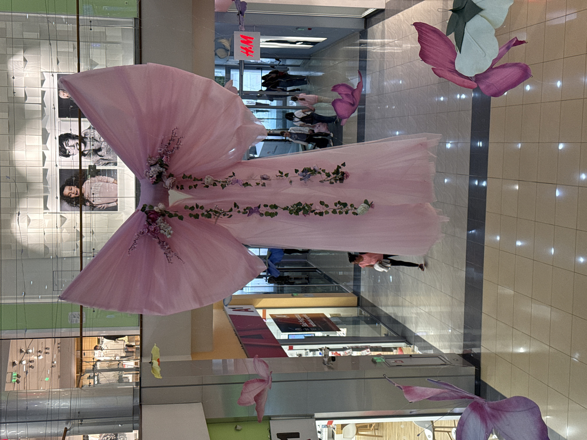 The image shows the interior of a shopping mall, possibly in Pleven, Bulgaria. A large pink tulle sculpture shaped like a dress or wings, adorned with floral decorations, is suspended from the ceiling. Several artificial pink and purple flowers are also hanging nearby. The mall features glass-fronted shops, including an H&M store visible through the glass. People can be seen walking through the mall. The floor is tiled with light-colored tiles, and the overall lighting is bright.