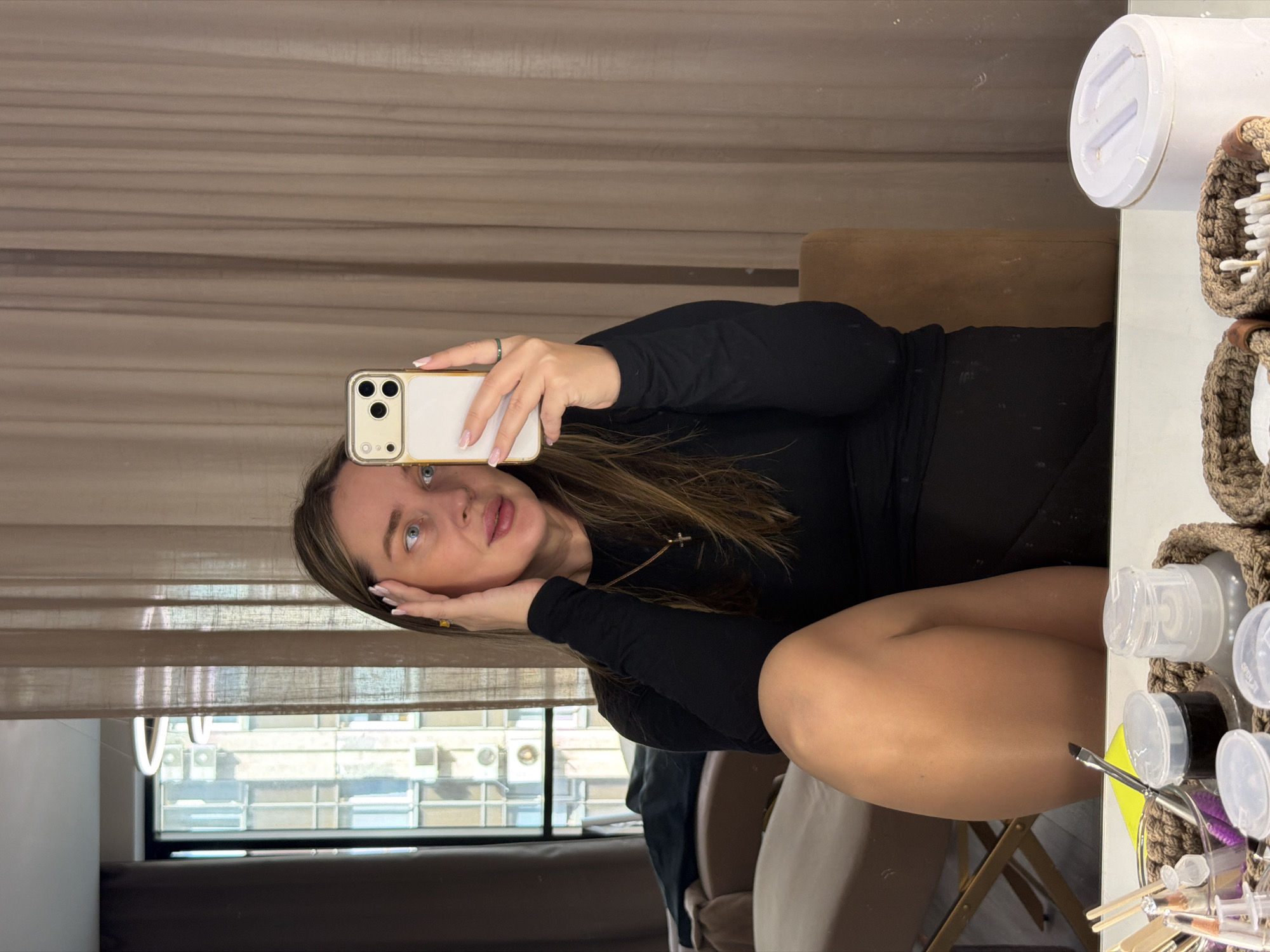 The image shows a woman taking a selfie in what appears to be a makeup or beauty setting. She is holding her phone with her left hand and has her right hand near her face. She is wearing a long-sleeved black outfit. The environment includes a mirror, makeup supplies in containers, and a window showing a cityscape with buildings. The city of Moscow is not confirmed by the elements in the scene, so that information is not included.
