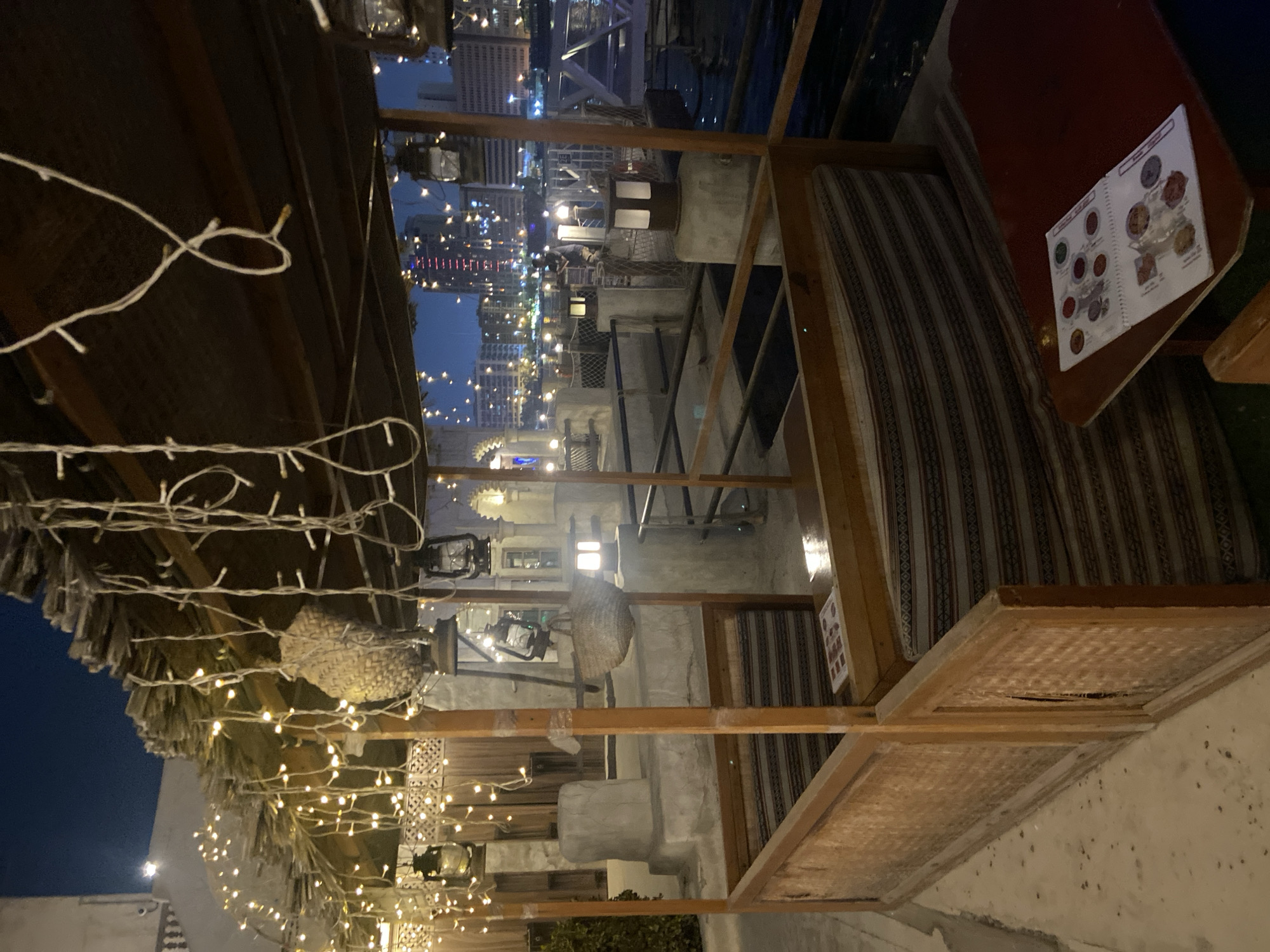 An outdoor dining area in Deira, United Arab Emirates, is captured at dusk. The area is covered by a thatched roof supported by wooden beams and adorned with strings of warm-toned fairy lights. Inside the seating area, cushioned benches with striped fabric offer seating. A dark red table in the foreground displays a menu with circular images of food items. In the background, a body of water, likely a creek, is visible, with a city skyline featuring illuminated buildings. Several vintage-style lanterns are also present in the scene, some hanging from the roof and others mounted on posts. The overall ambiance is cozy and illuminated by artificial light, with the twilight sky providing a dark blue backdrop.