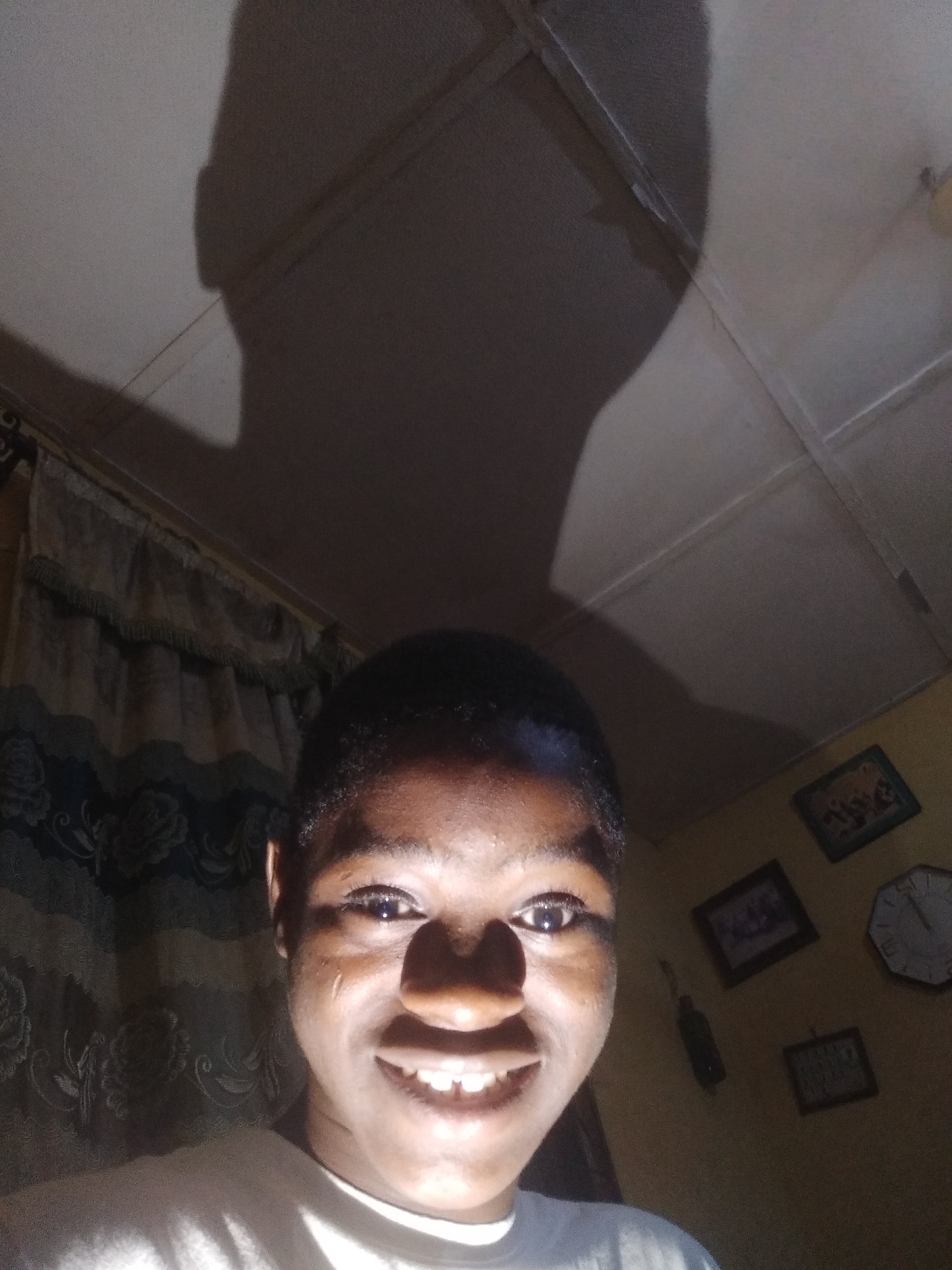 The image shows a person with short dark hair smiling into the camera. A shadow of their head is cast onto the ceiling behind them. The background consists of a curtain, a wall with framed pictures and a clock, and a textured ceiling. The location is inside a room. The city and state are unknown.