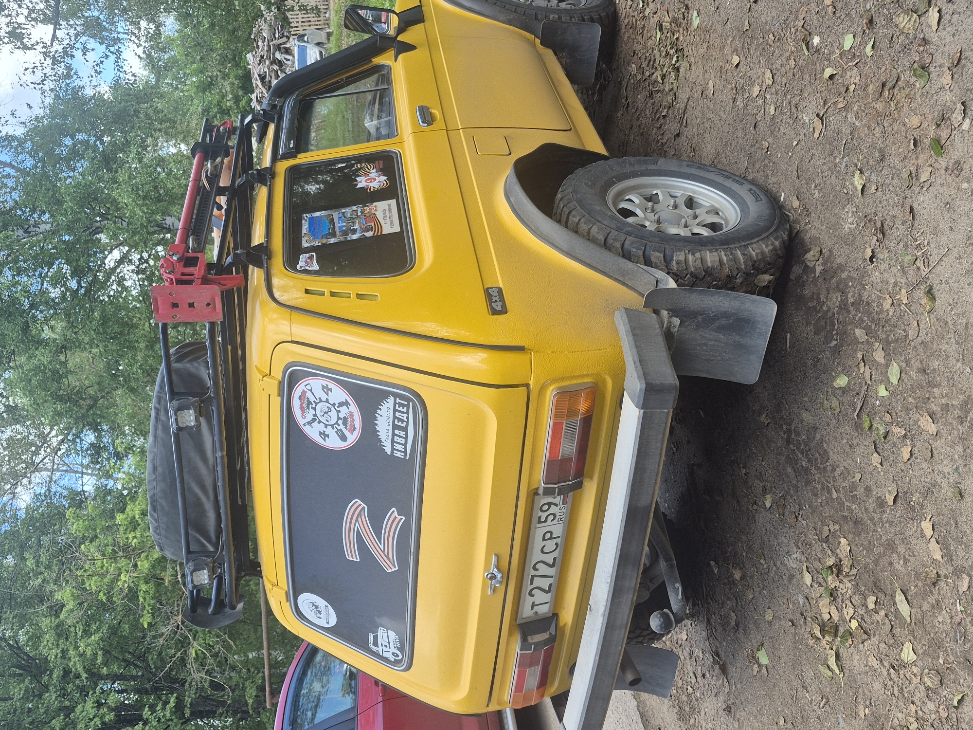 A yellow Lada Niva is parked on a dirt road in Chaikovsky, Russia. The car has a roof rack with a spare tire and a red high-lift jack. There are several stickers on the rear window, including a "Z" symbol and a sticker that reads "НИВА ЕДЕТ" which translates to "Niva is coming". The car is equipped with off-road tires and mud flaps. The environment is natural, with trees and foliage in the background. Fallen leaves are scattered on the ground.